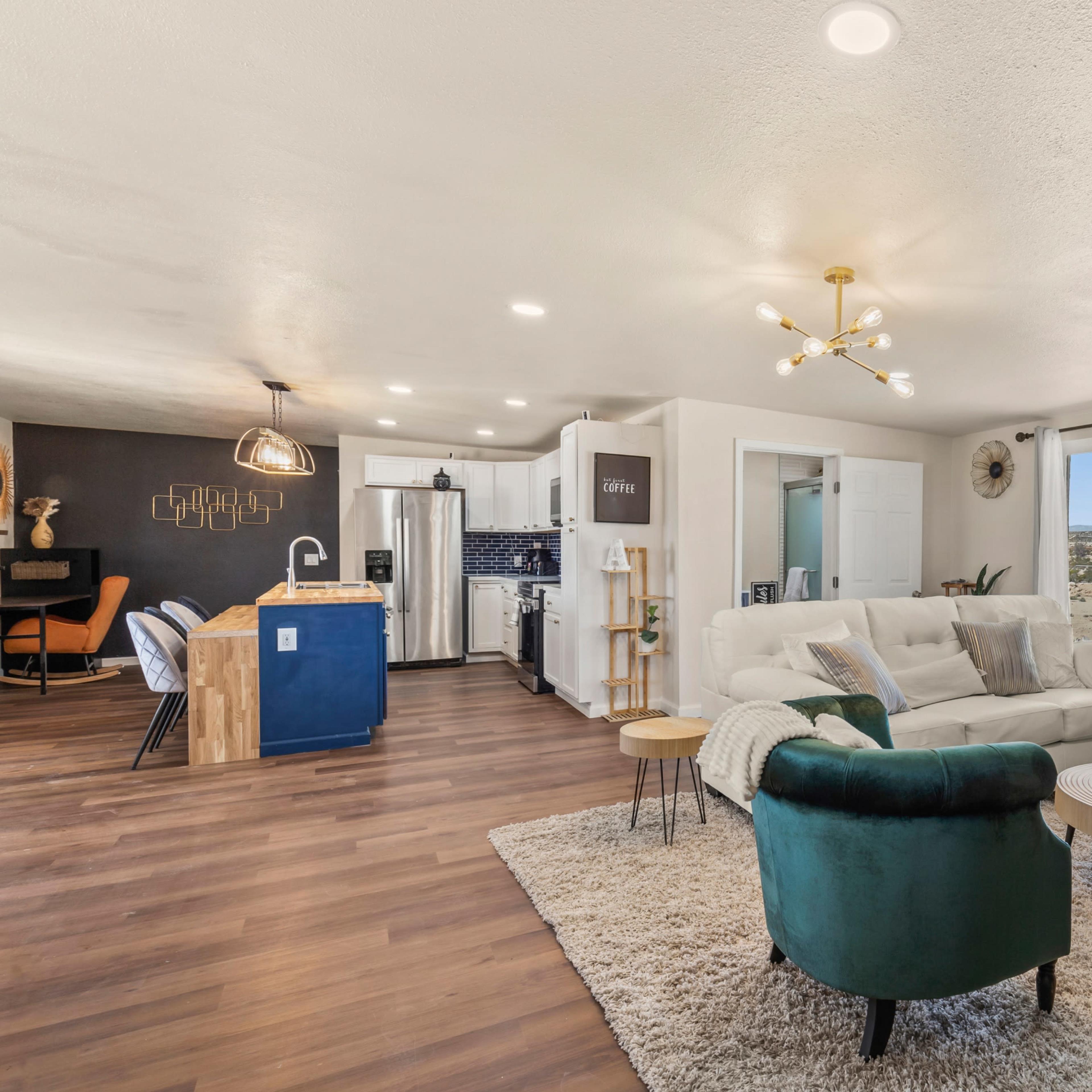 A modern living room features a white sofa, green accent chairs, a coffee table, and an open kitchen area with wooden and white cabinetry.