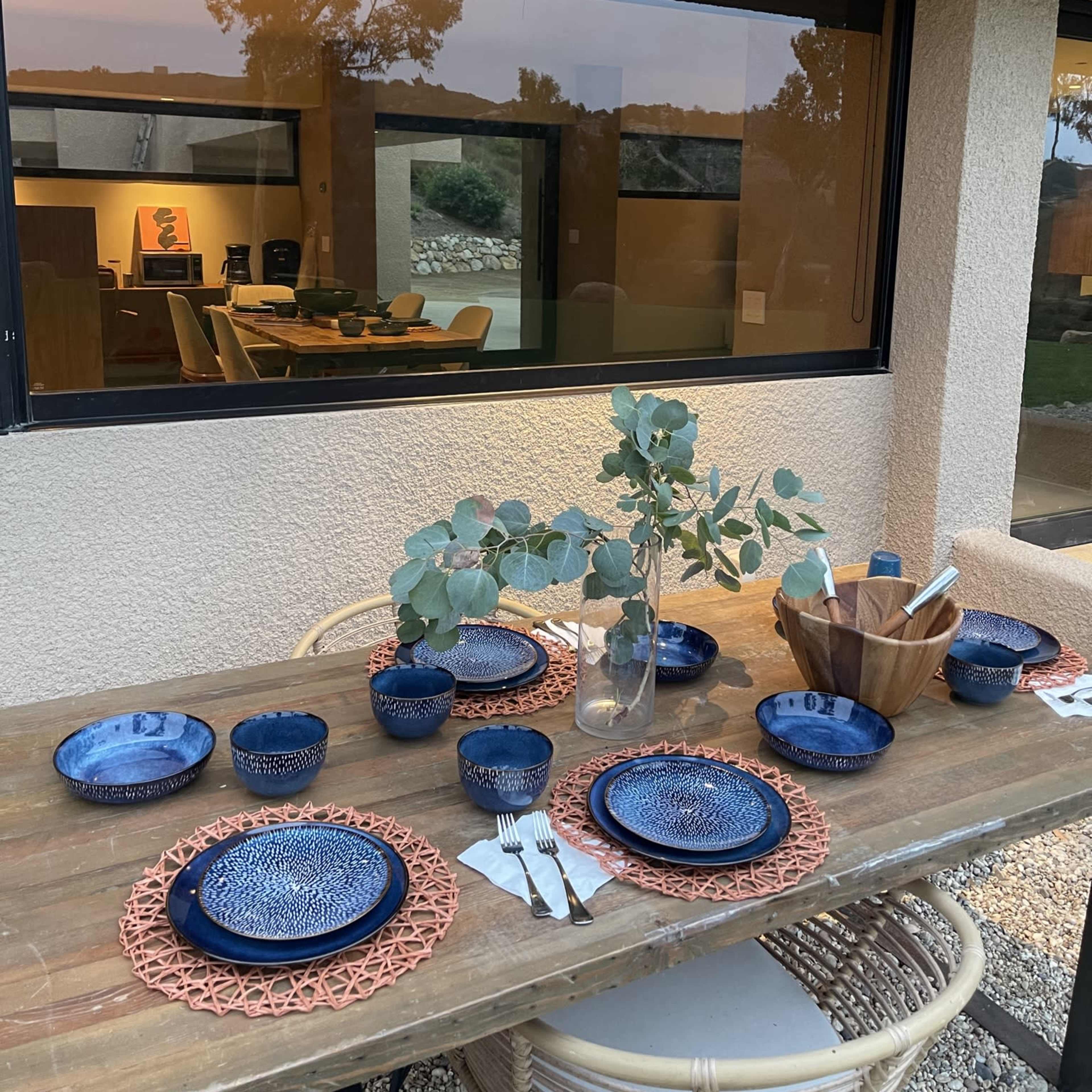 A dining table set with blue plates and a vase of eucalyptus sits outside next to a modern interior space visible through a large window.