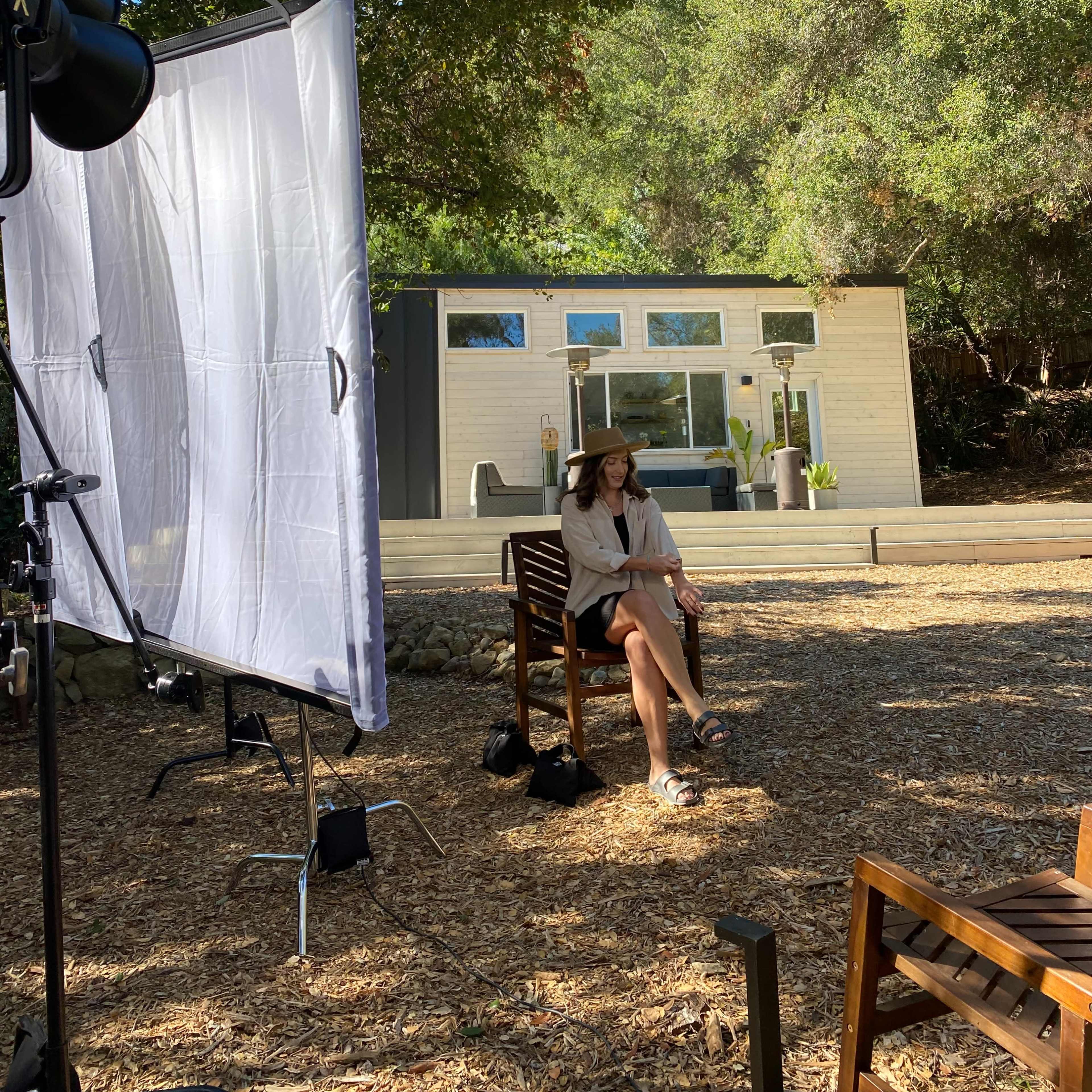 A woman sits on a wooden chair in a garden setting, with a modern tiny house in the background and photography equipment nearby.