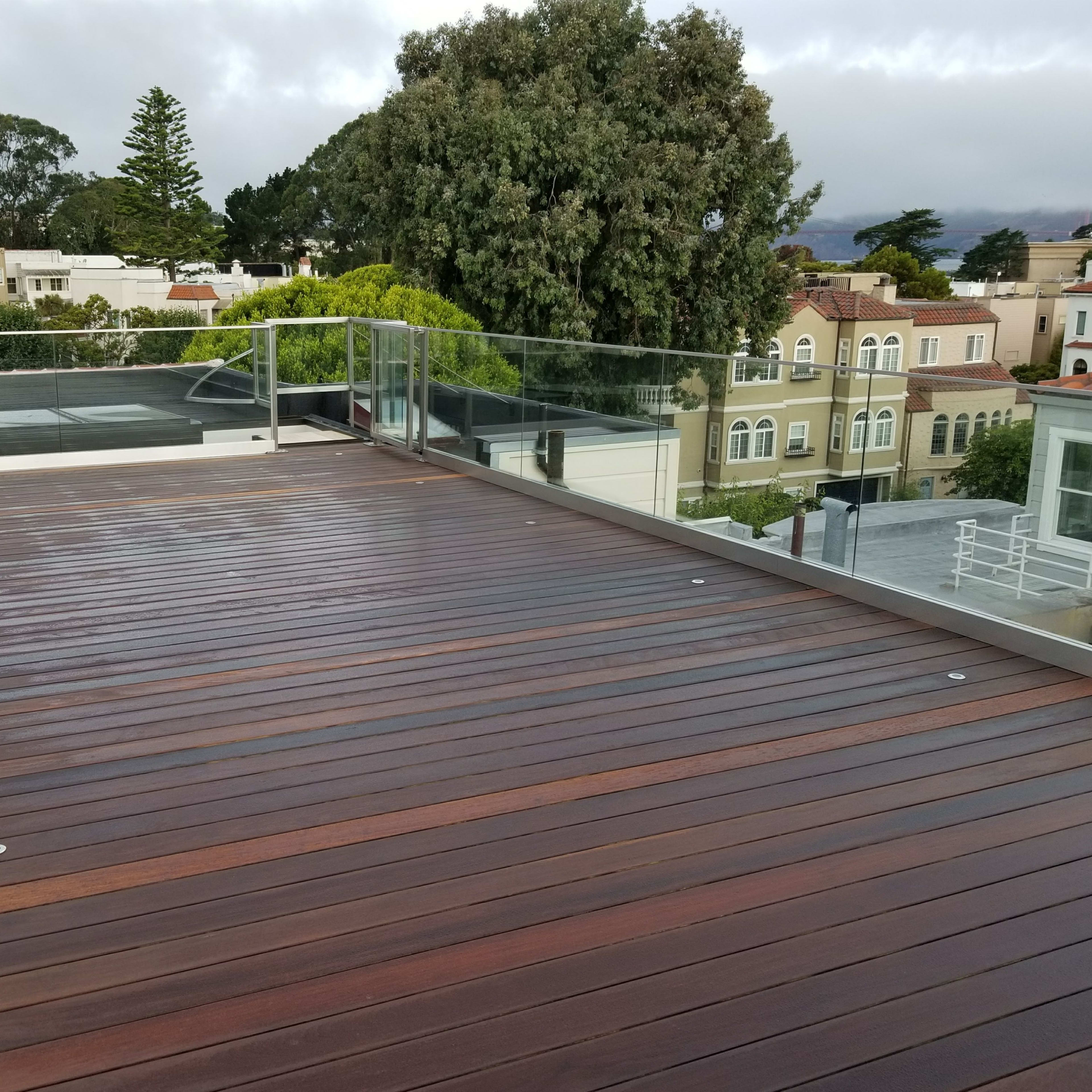 A wooden deck with a glass railing, overlooking a neighborhood of houses and greenery under a cloudy sky.