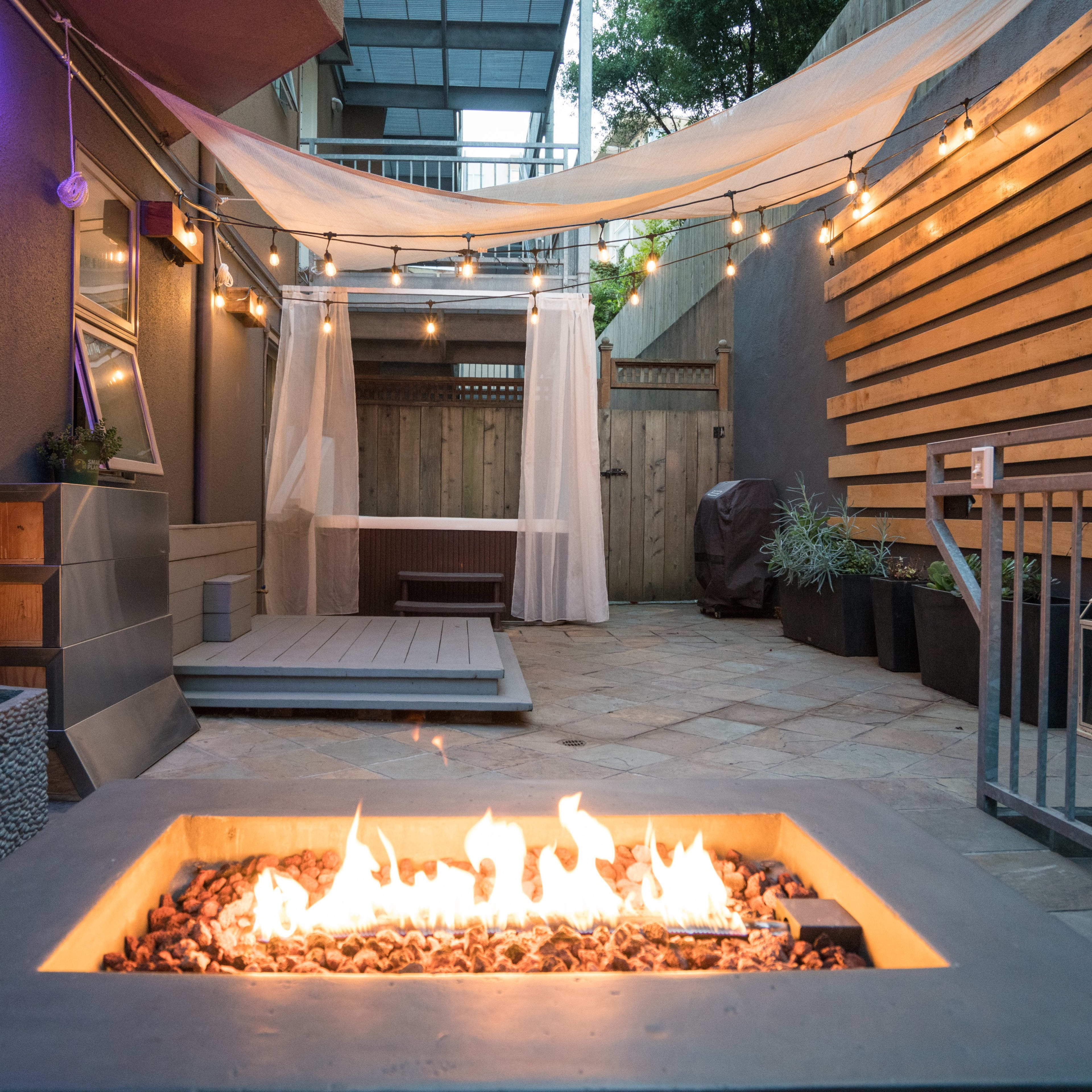 An outdoor patio area featuring a fire pit surrounded by stone and wooden elements, with string lights overhead and a shaded seating area in the background.
