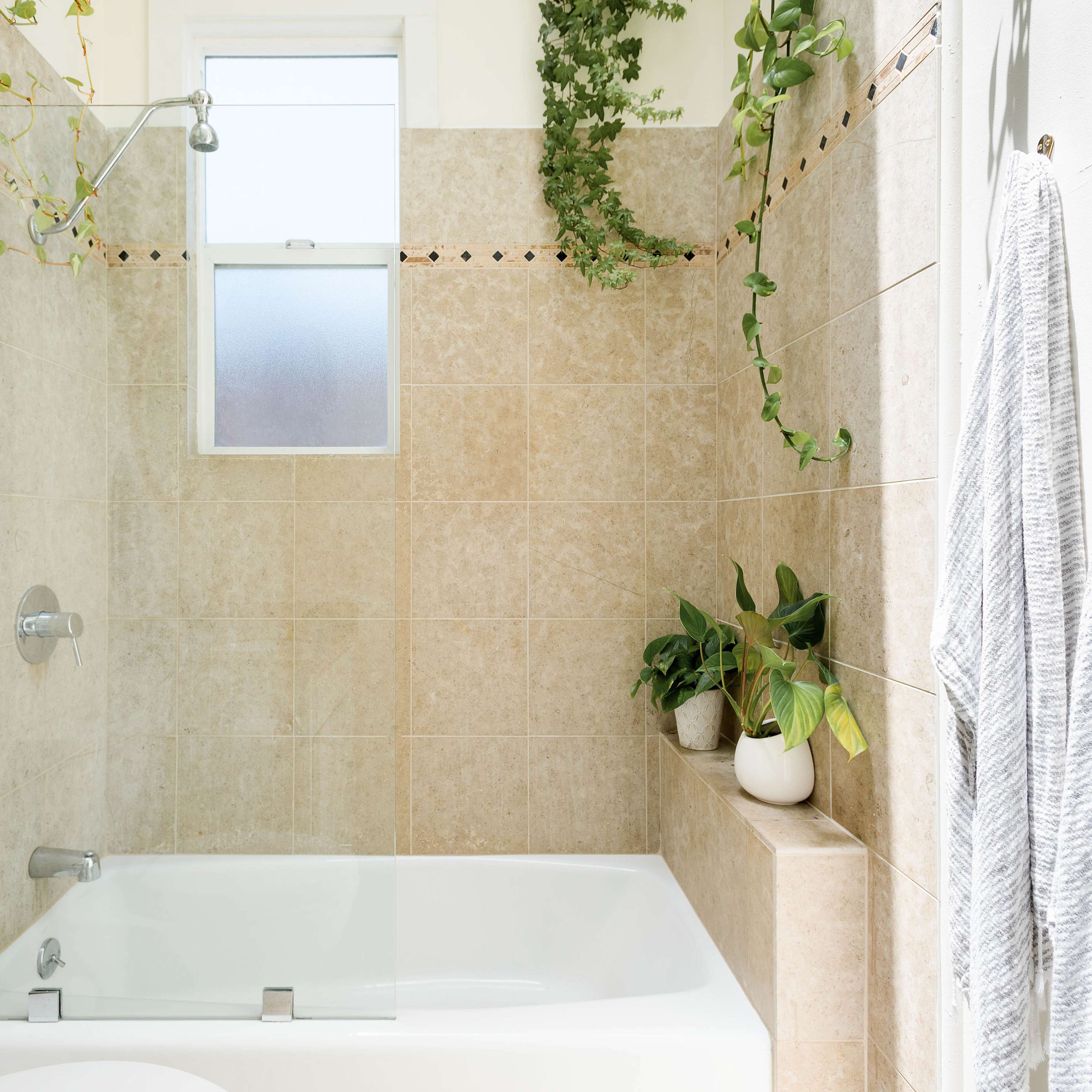 A bathroom featuring a bathtub with a shower, light-colored tile walls, and green plants positioned around the area.