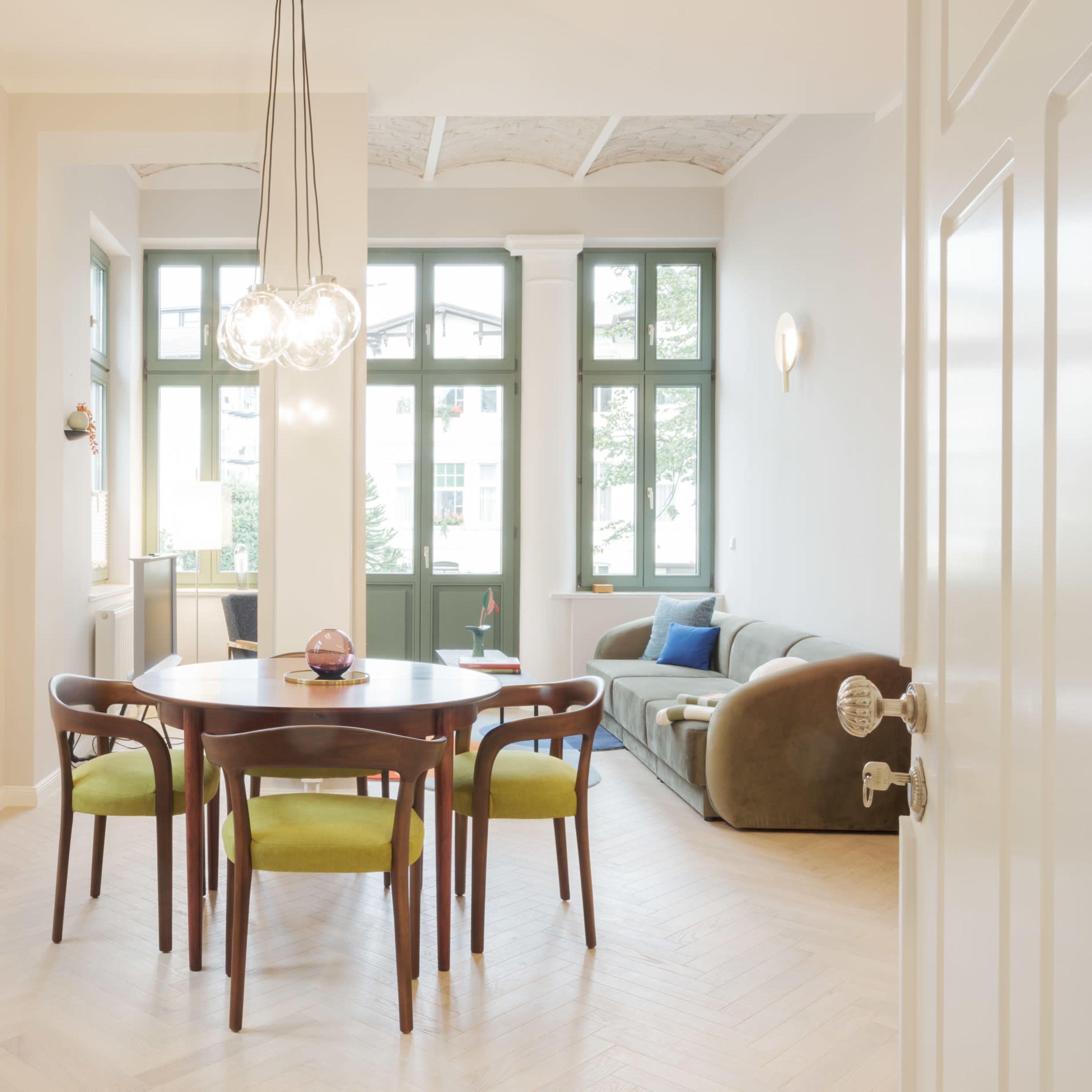A dining area with a round wooden table surrounded by chairs, set against a backdrop of large green-framed windows and a cozy living space featuring a sofa.