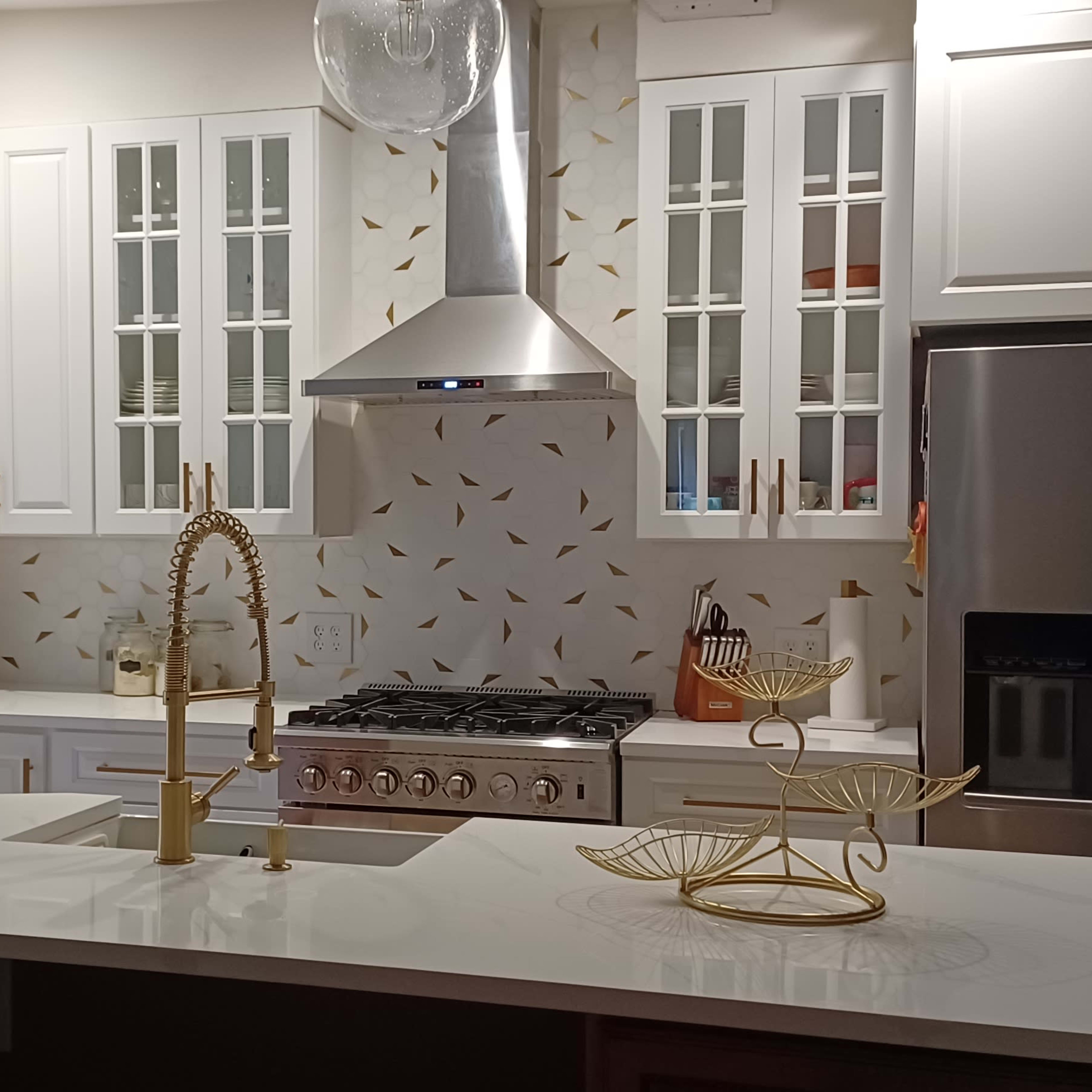 A modern kitchen features white cabinetry, a stainless steel range hood, and a large island with a gold-accented sink.