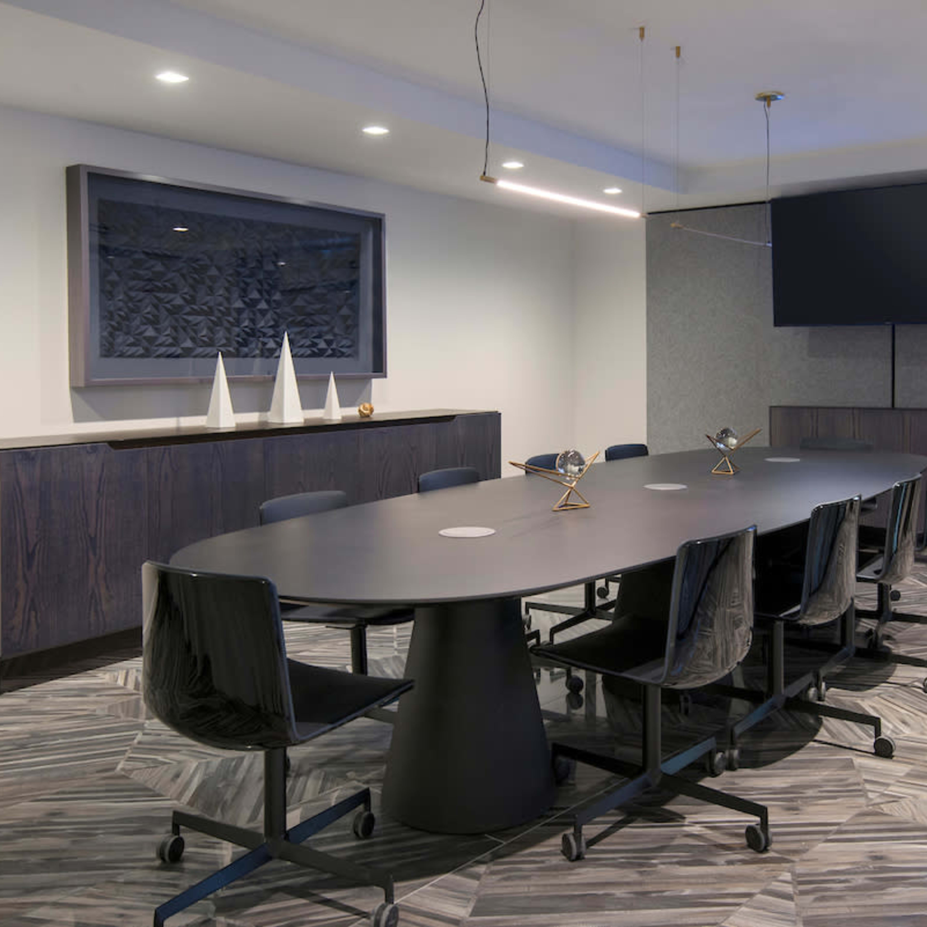A long, oval conference table is surrounded by rolling black chairs in a modern meeting room featuring a large screen and decorative wall art.