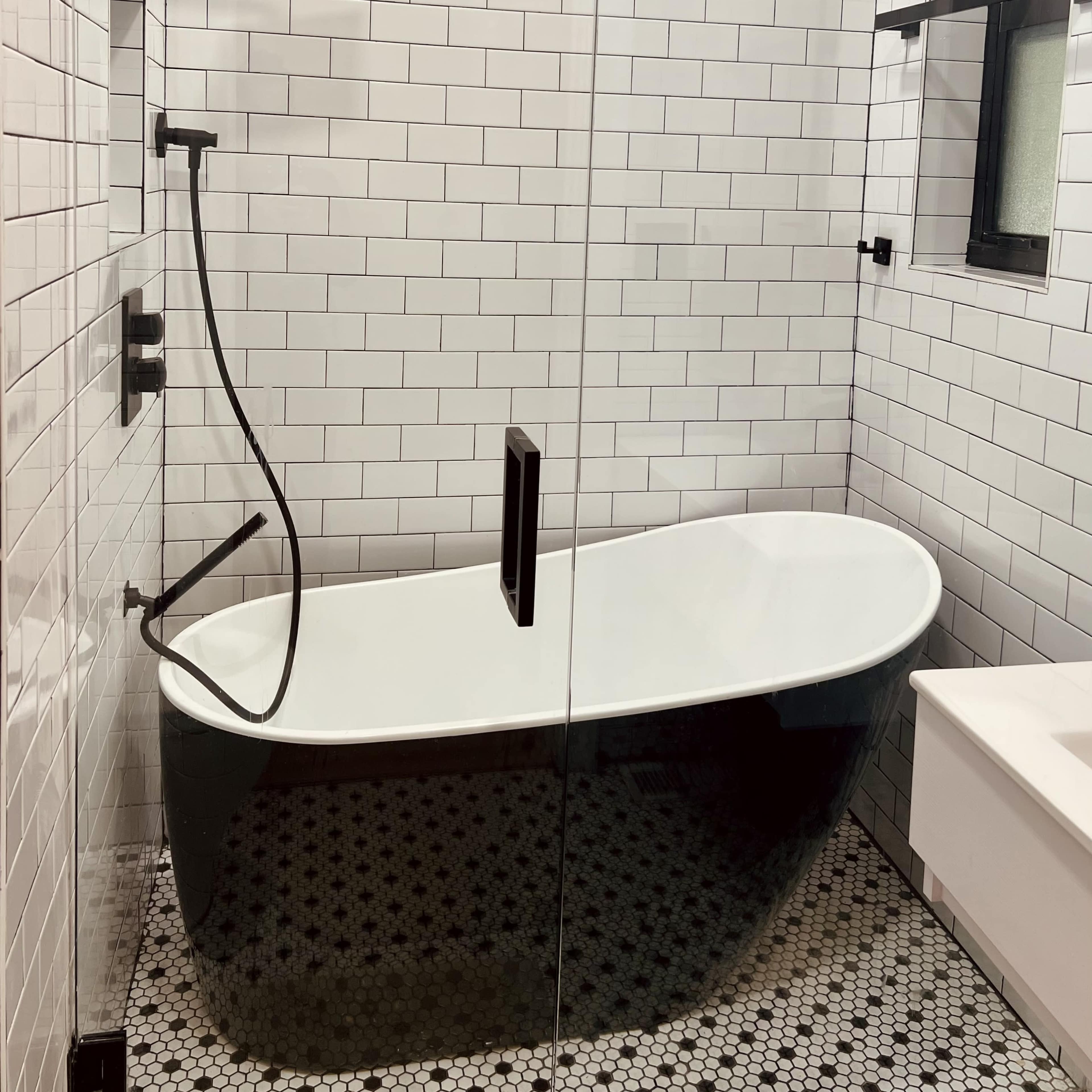 A modern bathroom featuring a freestanding black and white bathtub, a glass shower enclosure, and white subway tile walls.