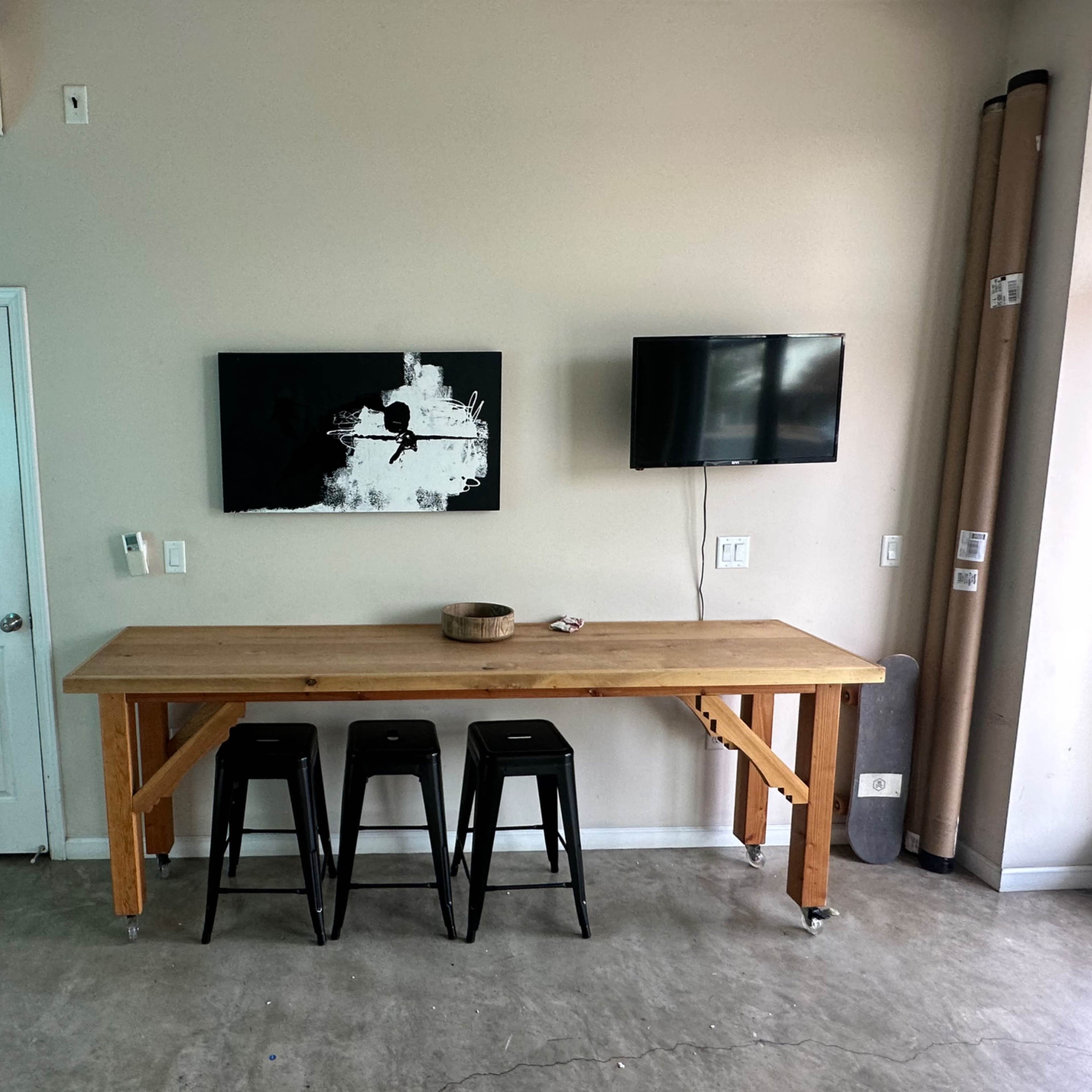 A wooden table with three black stools stands against a wall, featuring two televisions and a piece of artwork above it.