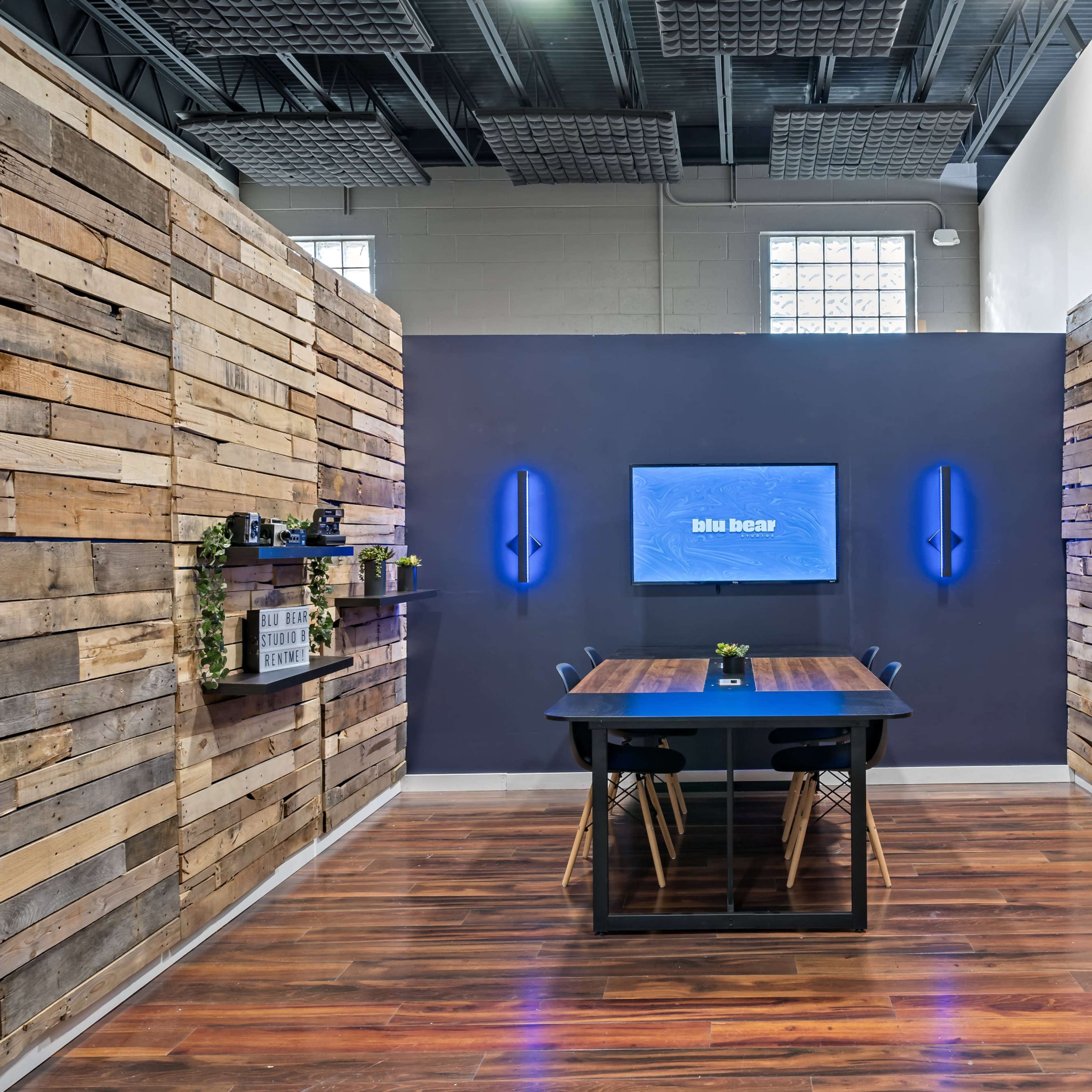 A modern meeting room with wooden plank walls, a dark table surrounded by chairs, and a wall-mounted screen displaying a blue logo.