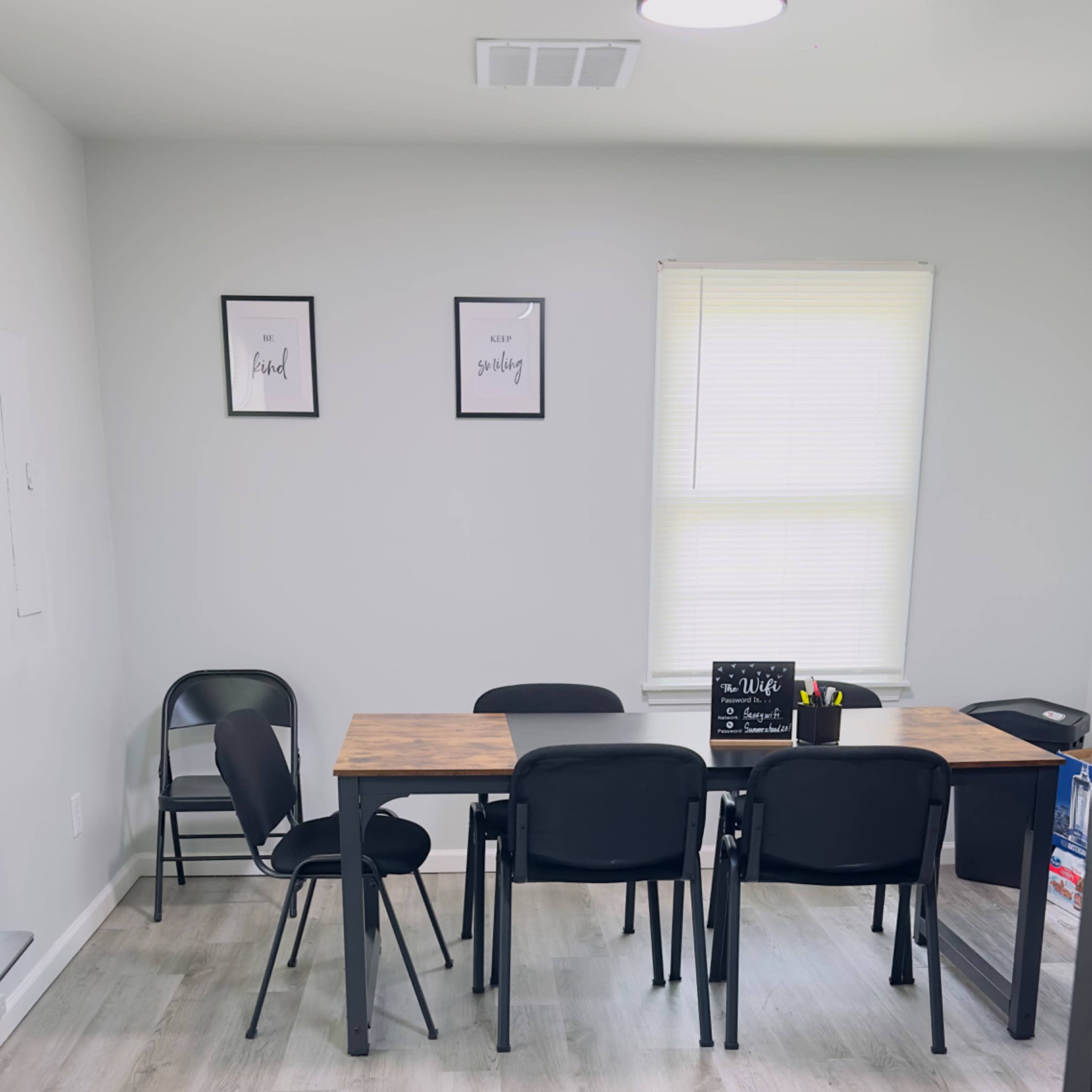 A simple meeting room features a wooden table surrounded by black chairs, with framed artwork on the walls and a window letting in natural light.