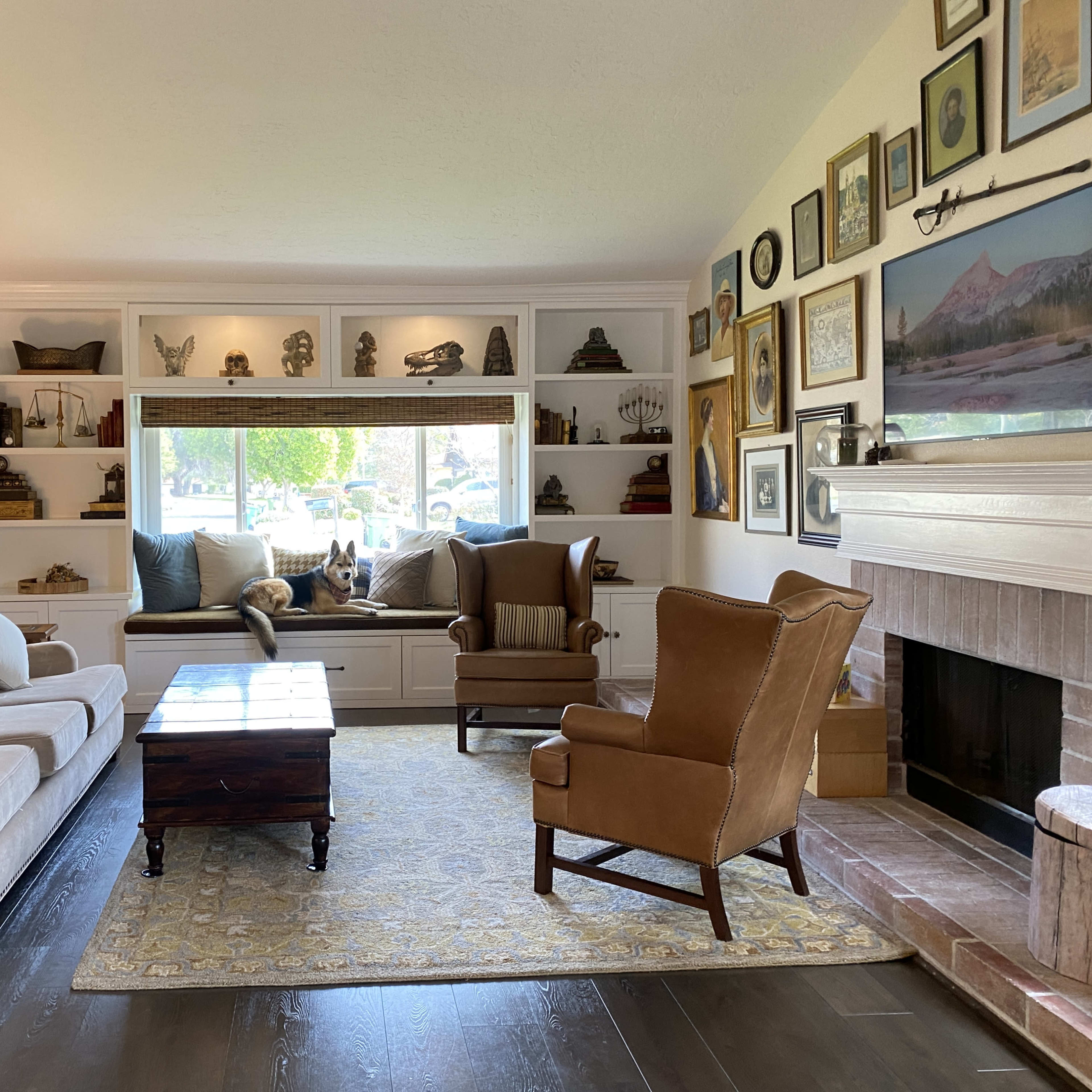 A living room features a beige sofa, a wooden coffee table, and an armchair, with built-in shelves displaying various decorative items and a window seat in the background.