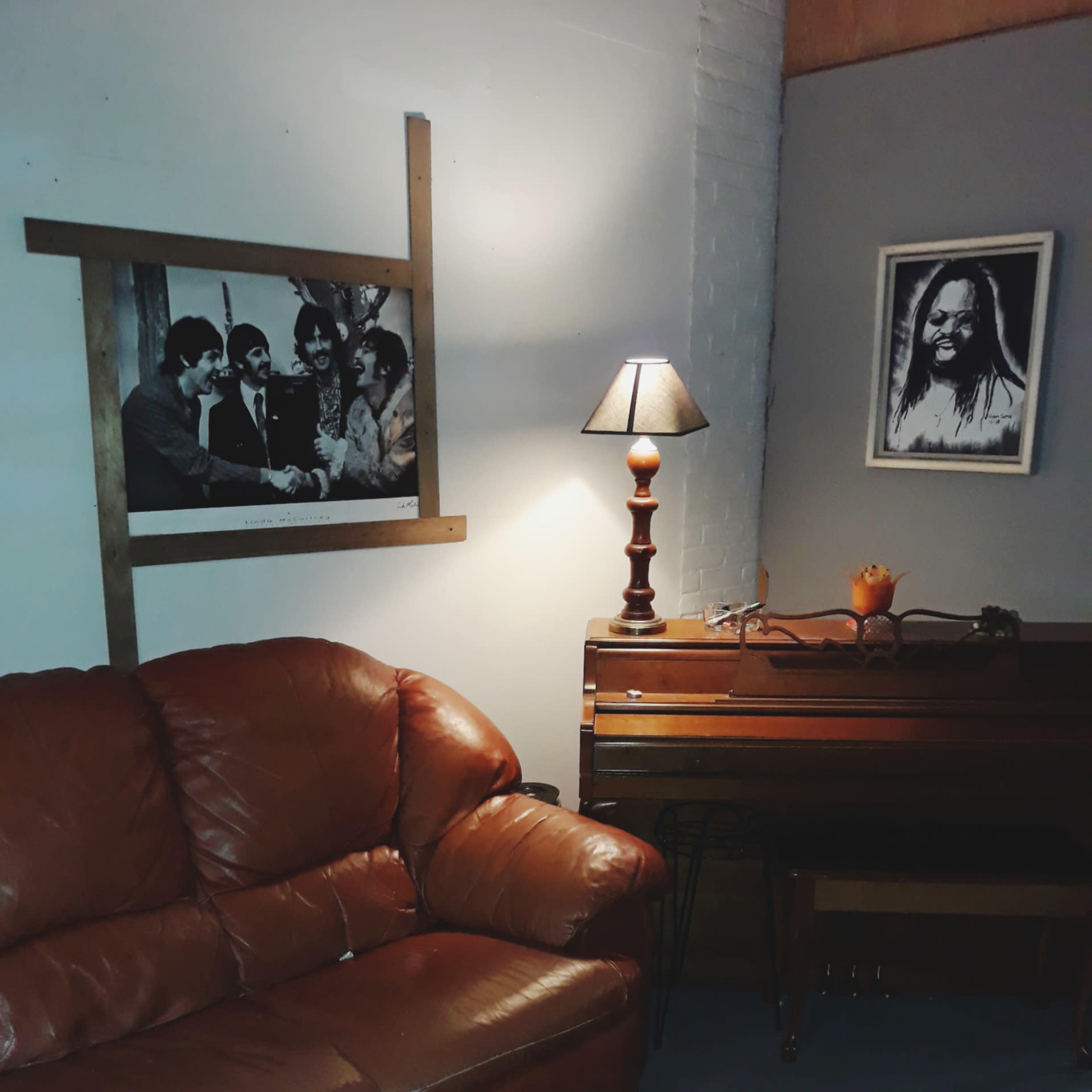 A modest living room with a brown leather couch, a lamp, a wooden piano, and framed photographs on the walls.