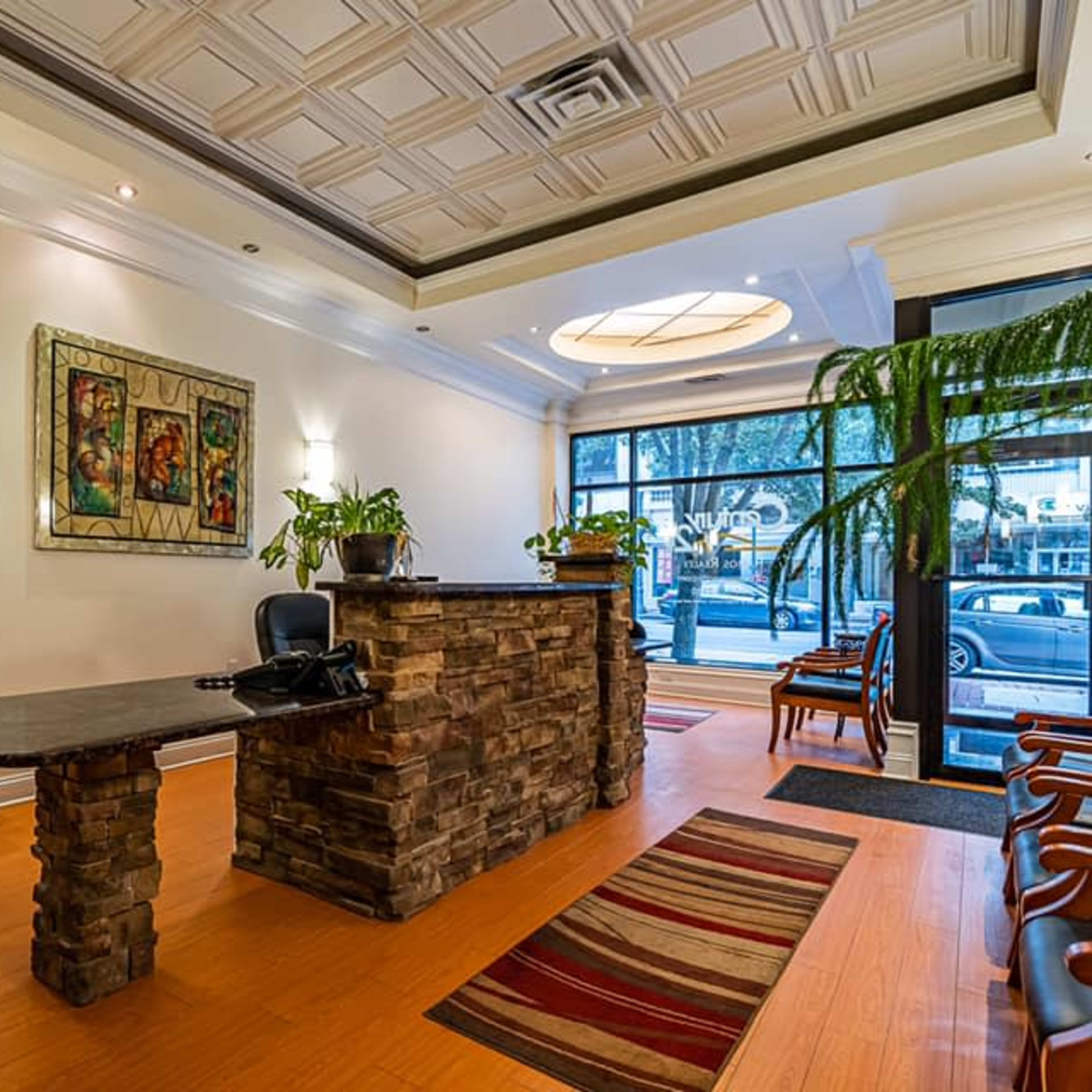 A modern reception area with a stone front desk, a printer, a water cooler, and potted plants, designed with wooden flooring and large windows.