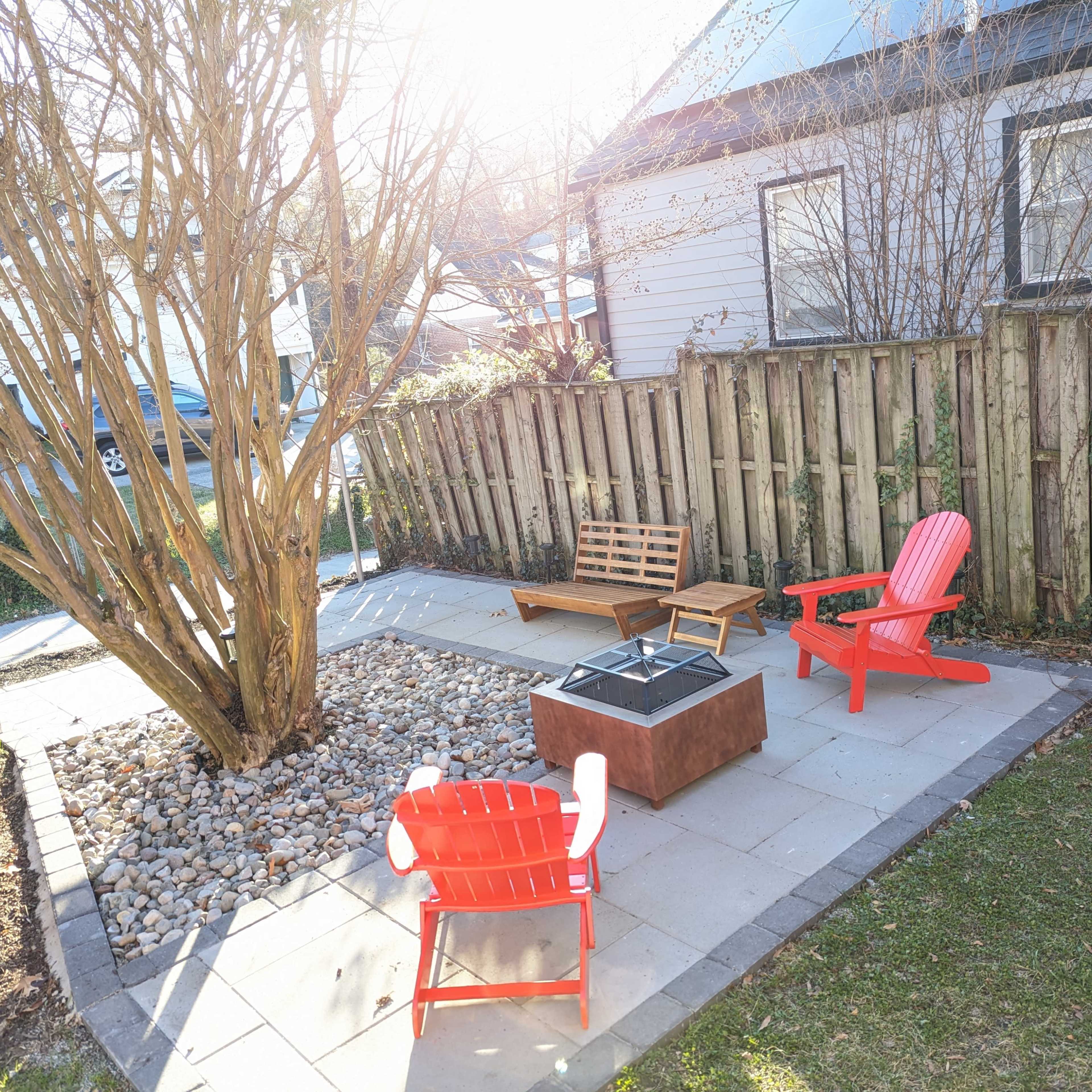 A small outdoor patio with four chairs and a fire pit surrounded by stones and a tree, enclosed by wooden fencing.