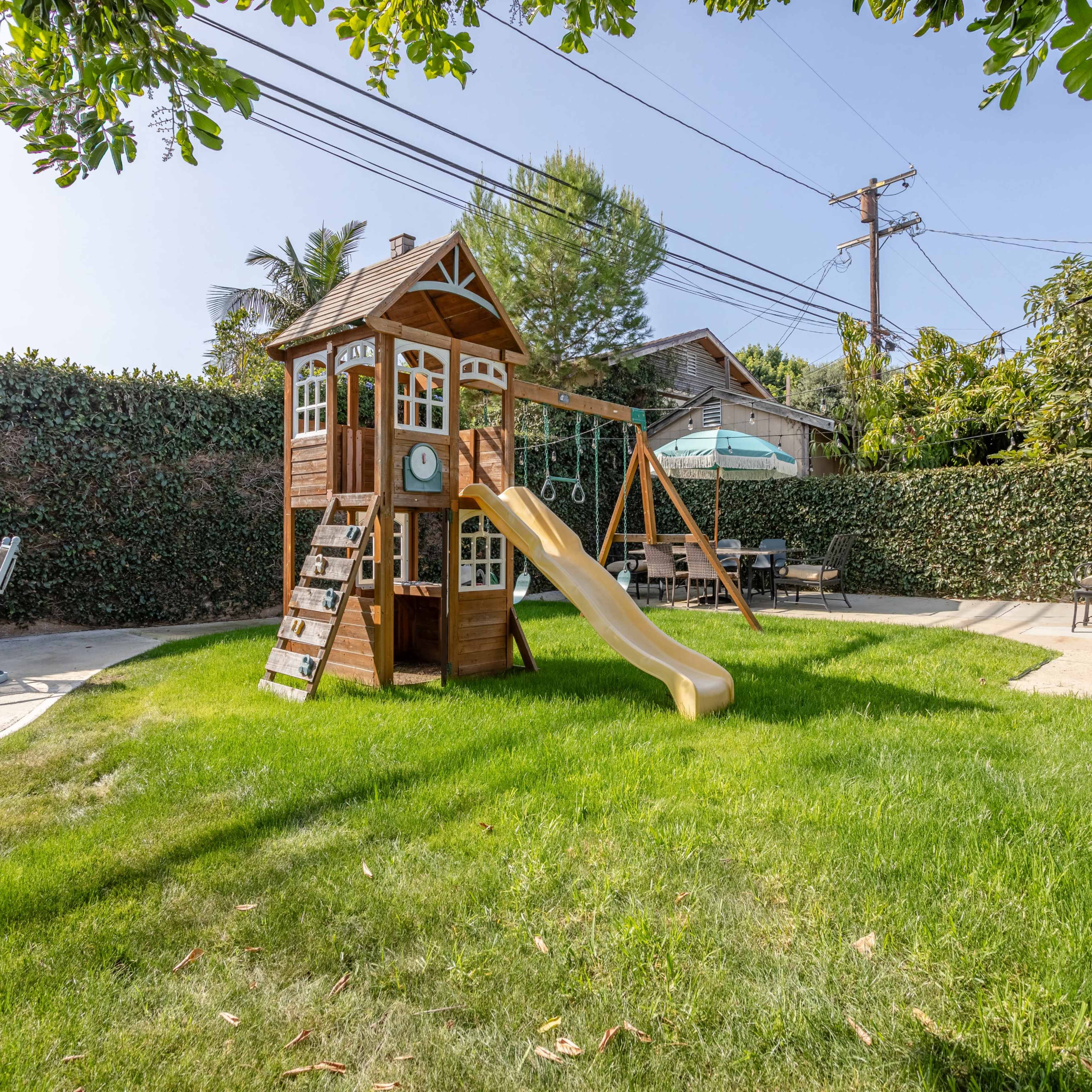 A wooden play structure with a slide and swing set is located on a grassy area surrounded by hedges in a backyard.