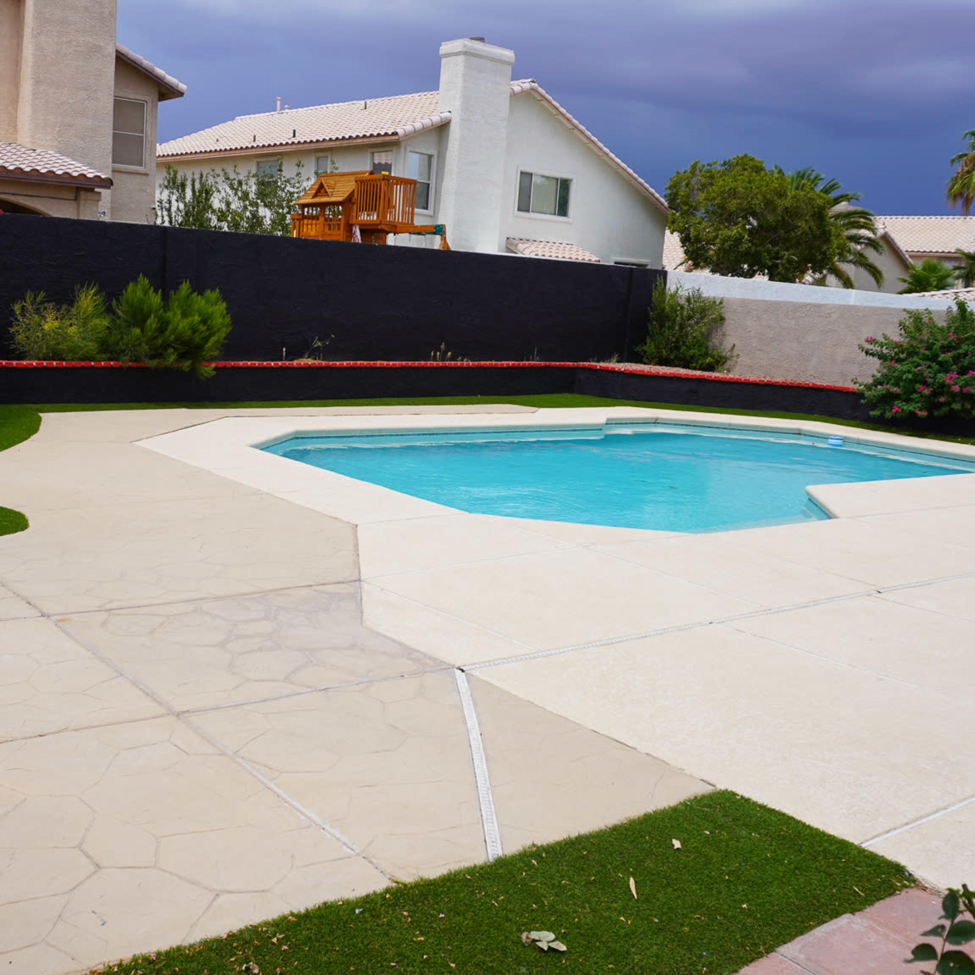 A backyard with a swimming pool surrounded by concrete and a grassy area, featuring a palm tree and a playset in the background.