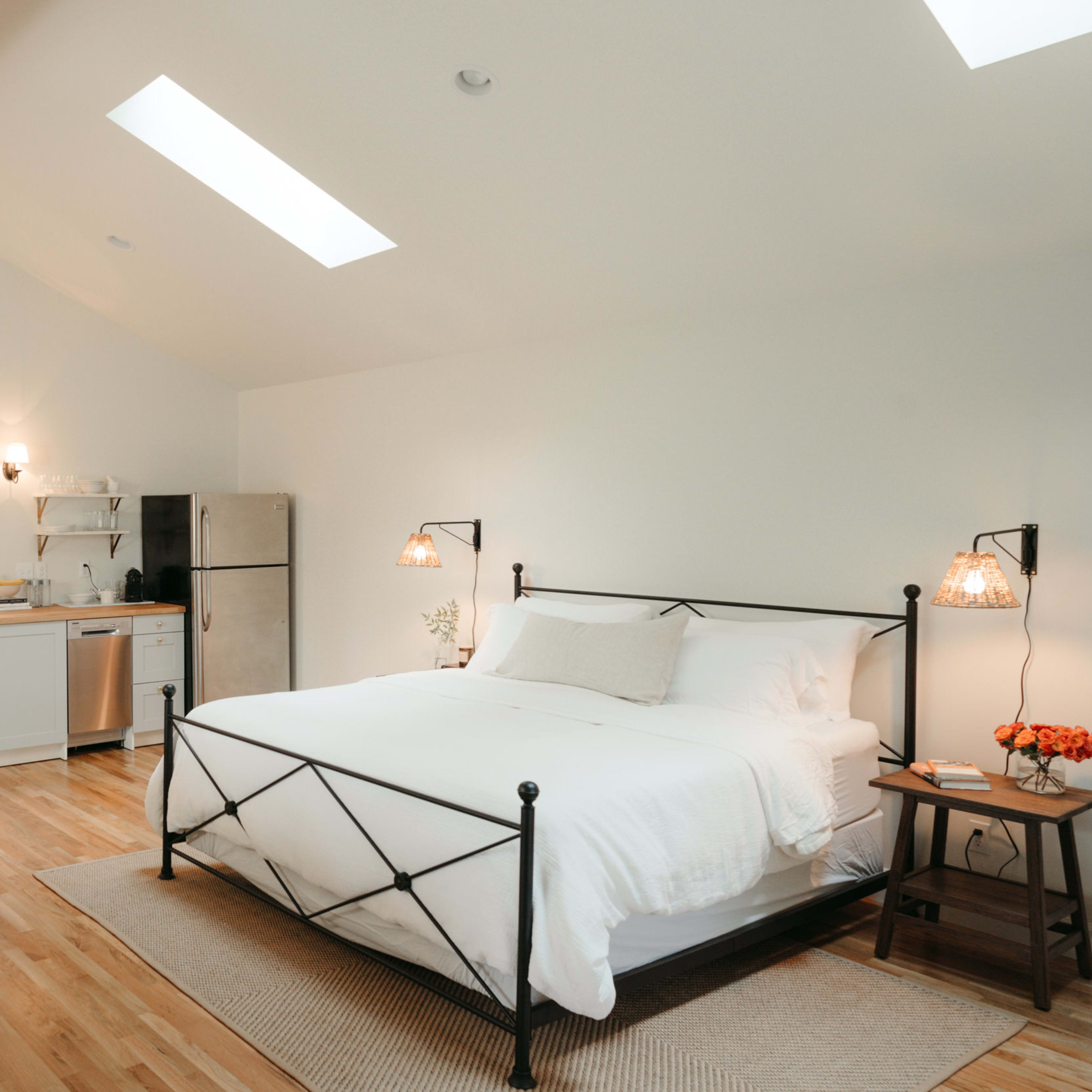 A bright and airy bedroom with a large bed, a kitchenette, and a sitting area, featuring wooden beams and large windows.