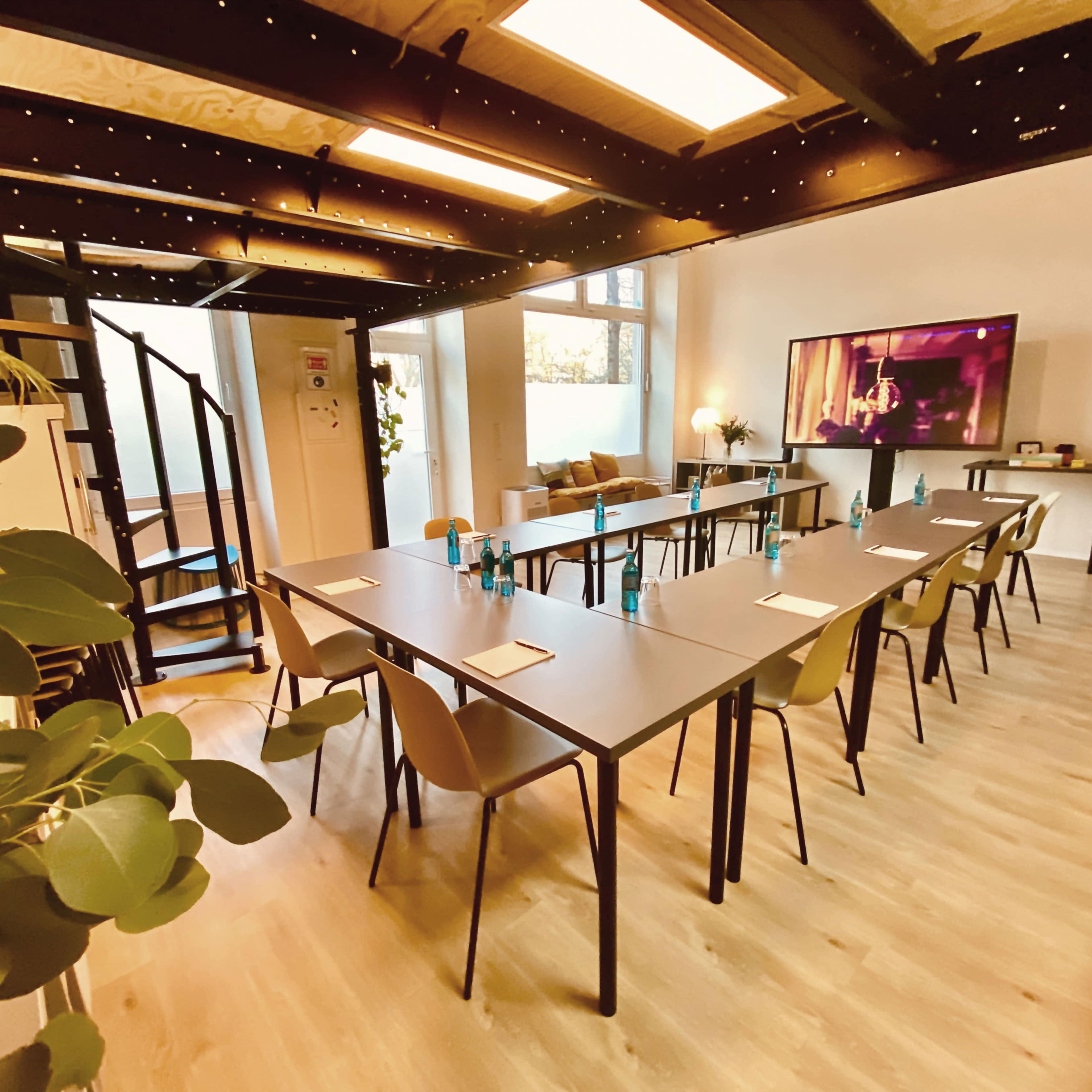 A modern meeting room features a set of long tables arranged for a conference, with chairs and a large screen at one end.