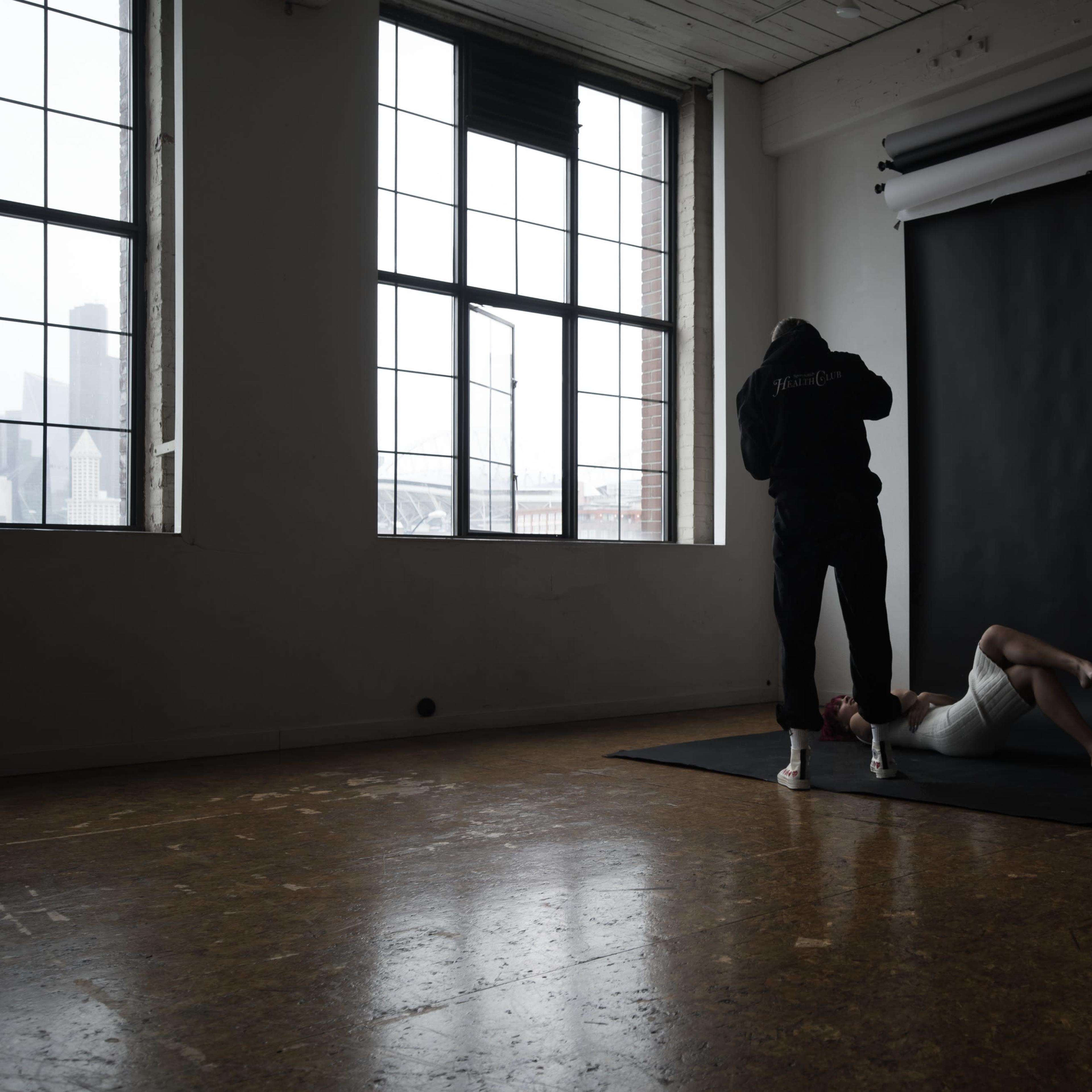 A person in a dark outfit is positioning a model lying on a black mat in a sparsely furnished studio with large windows.