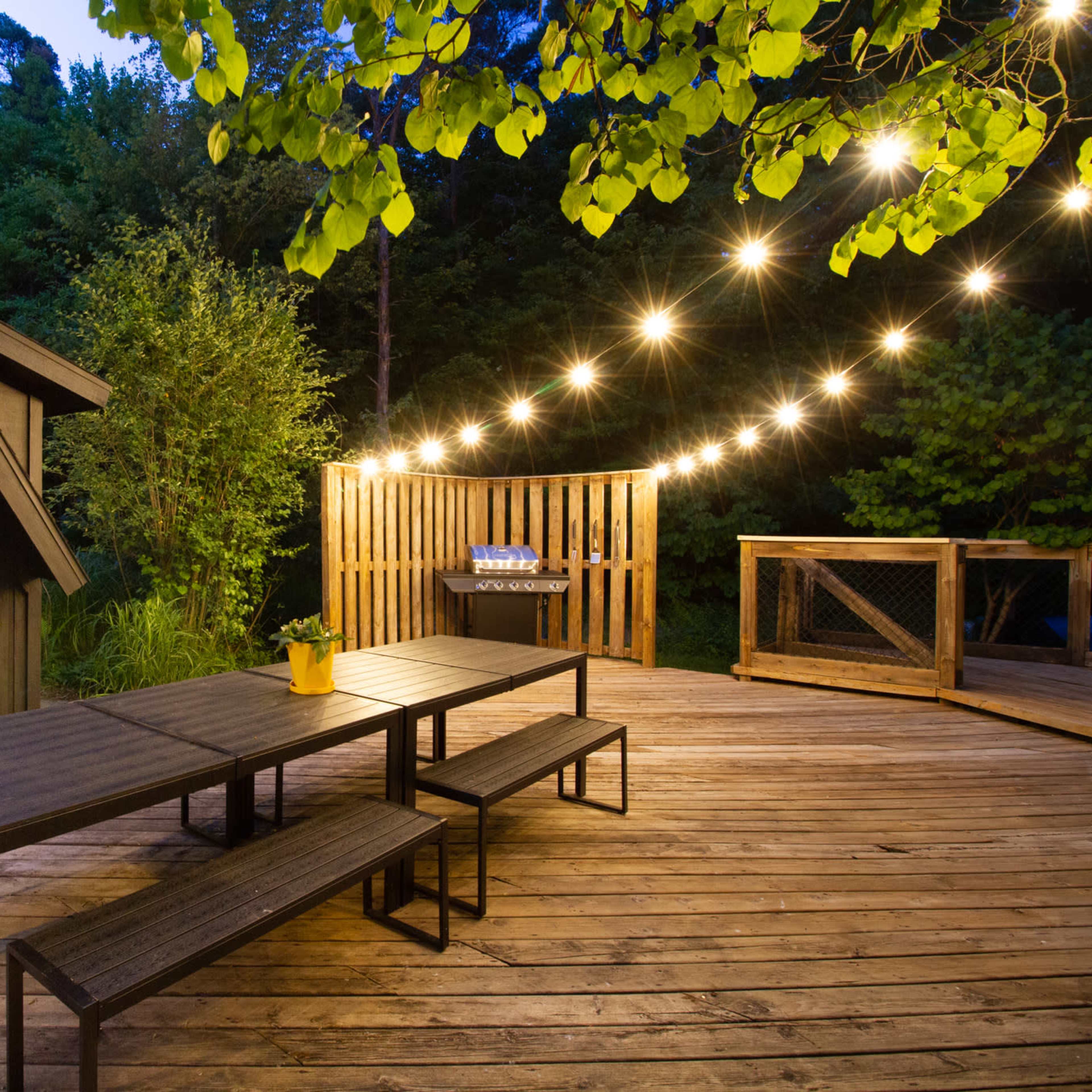 A wooden deck with a large table and benches is illuminated by string lights, adjacent to a house with a large window surrounded by greenery.