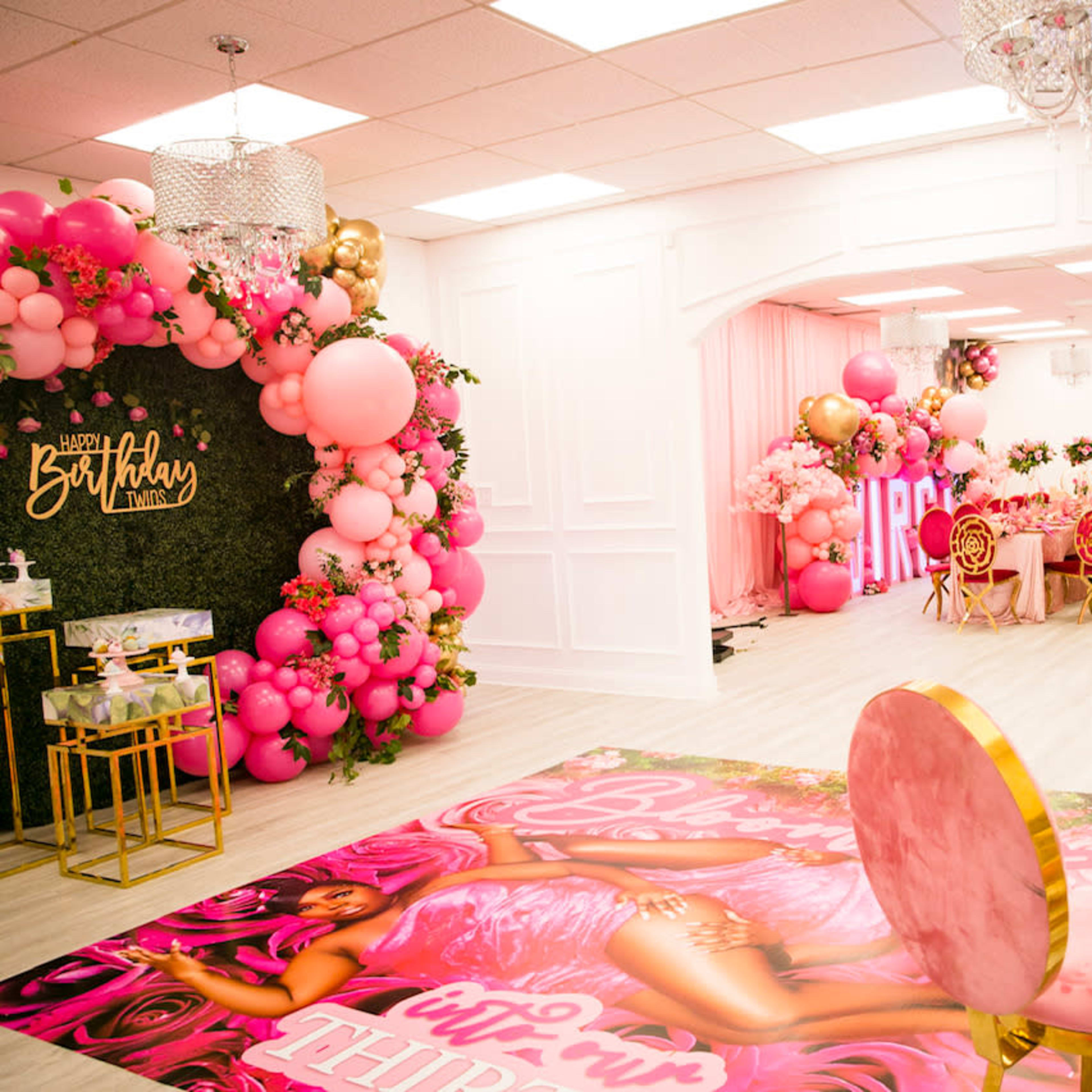 A decorated event space featuring pink balloon arrangements, a lush floral backdrop, and a themed floor mat with a seating area of gold chairs and a table.
