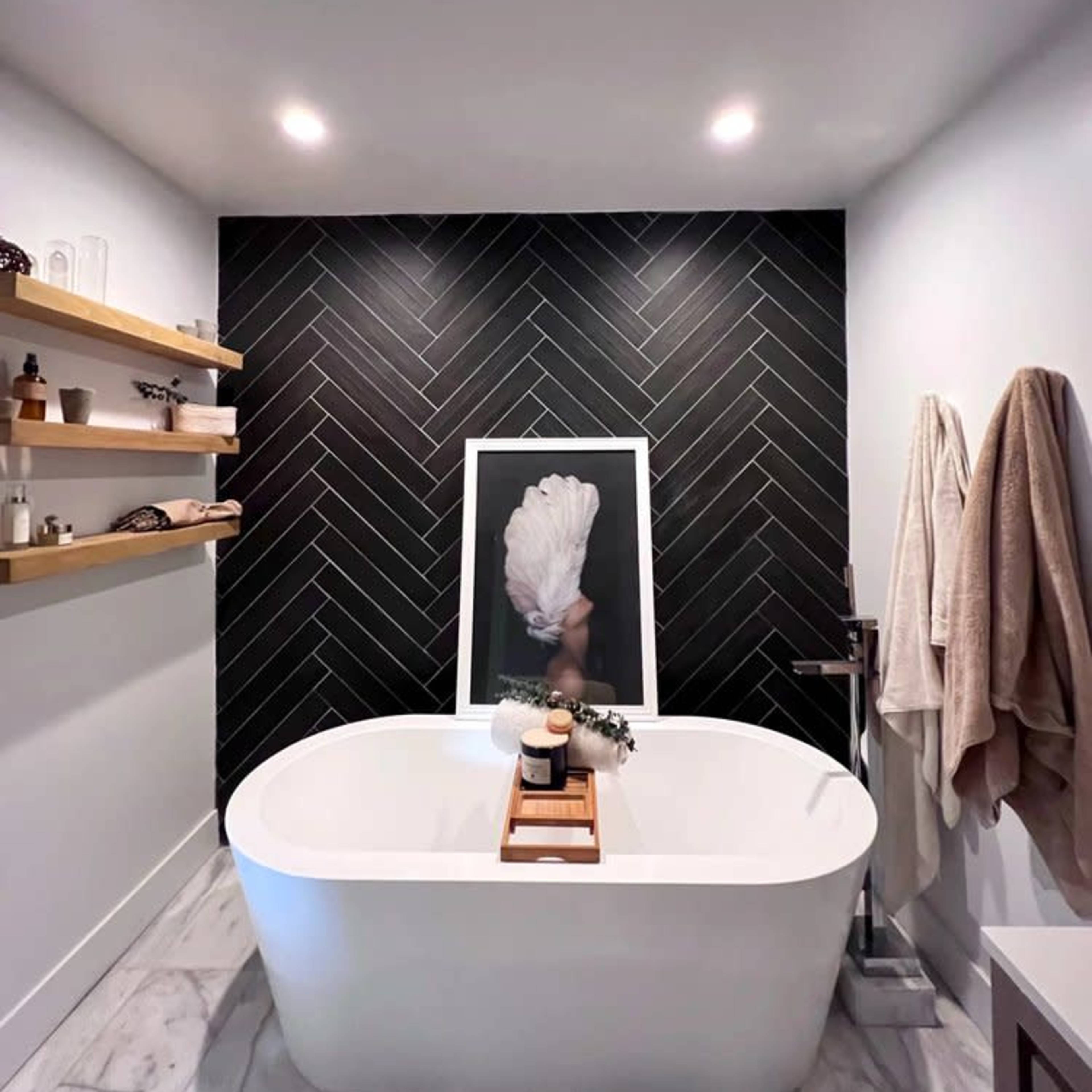 A modern bathroom featuring a freestanding white bathtub, a dark herringbone tile accent wall, wooden floating shelves, and a towel hanging to the side.