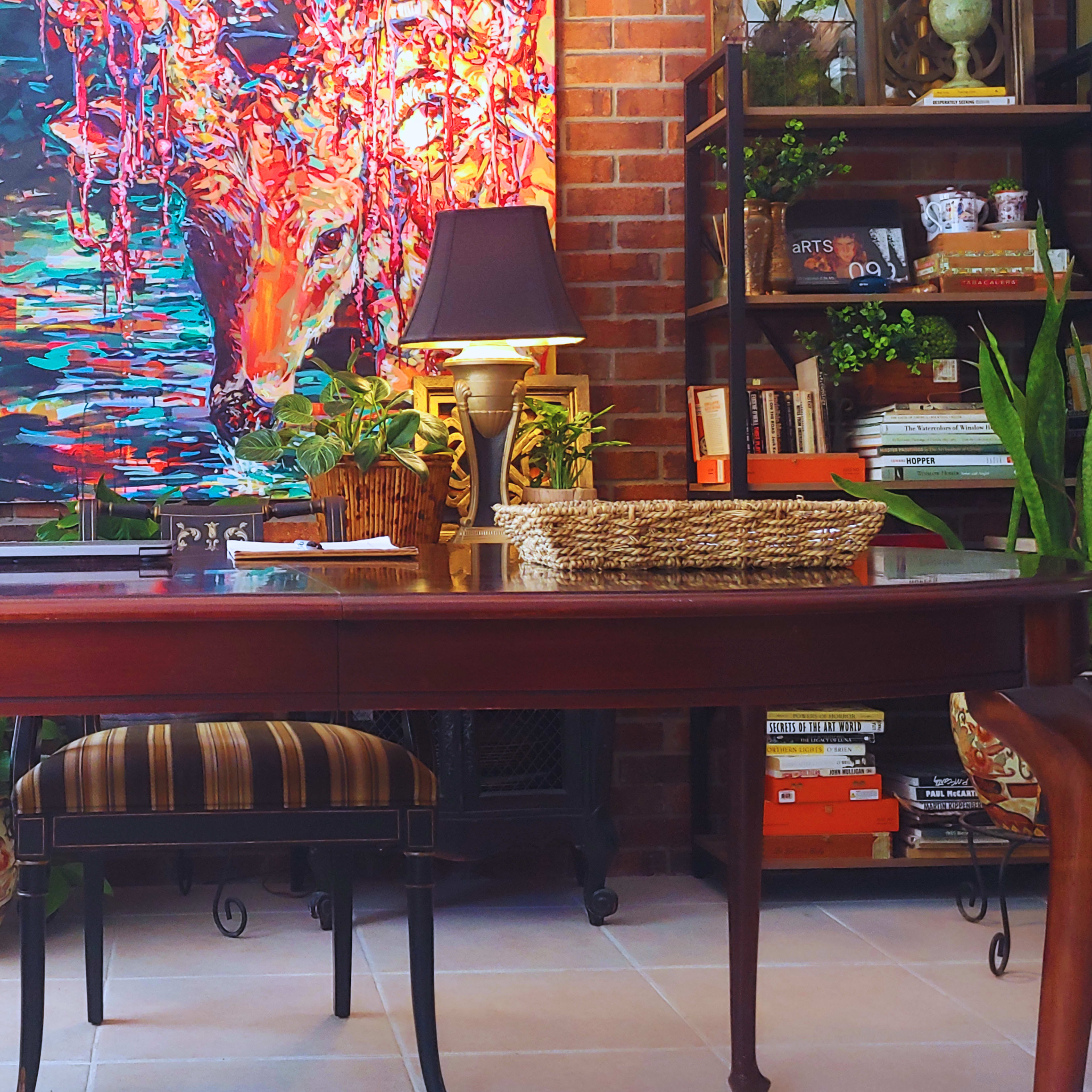 A wooden desk is positioned in front of a colorful abstract painting, surrounded by plants and bookshelves filled with books and decorative items.