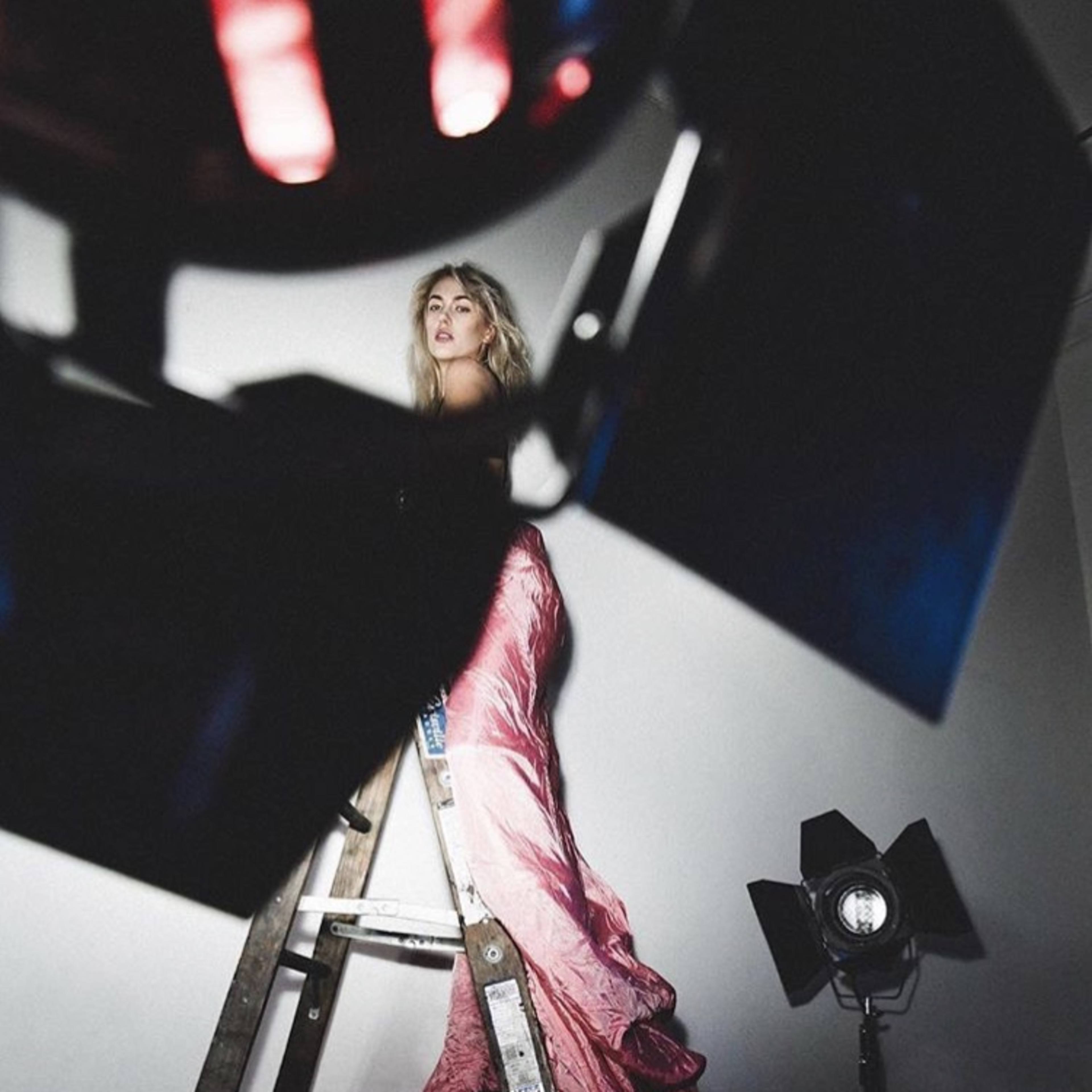 A model poses on a ladder while wearing a long, flowing garment, with studio lights illuminating the scene.