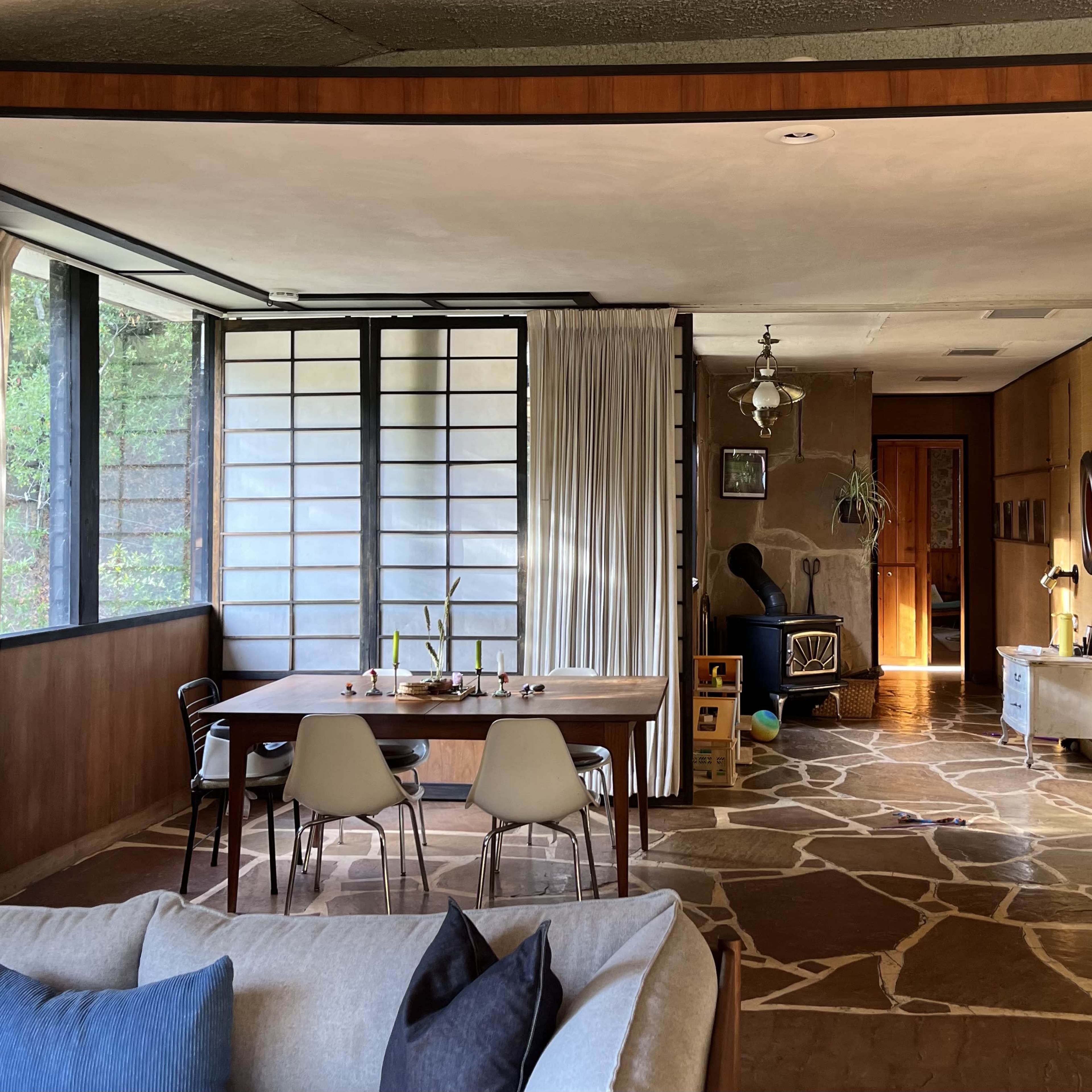 A spacious, modern living area with a dining table, chairs, and a mix of natural light and shadow from large windows and an adjacent room.