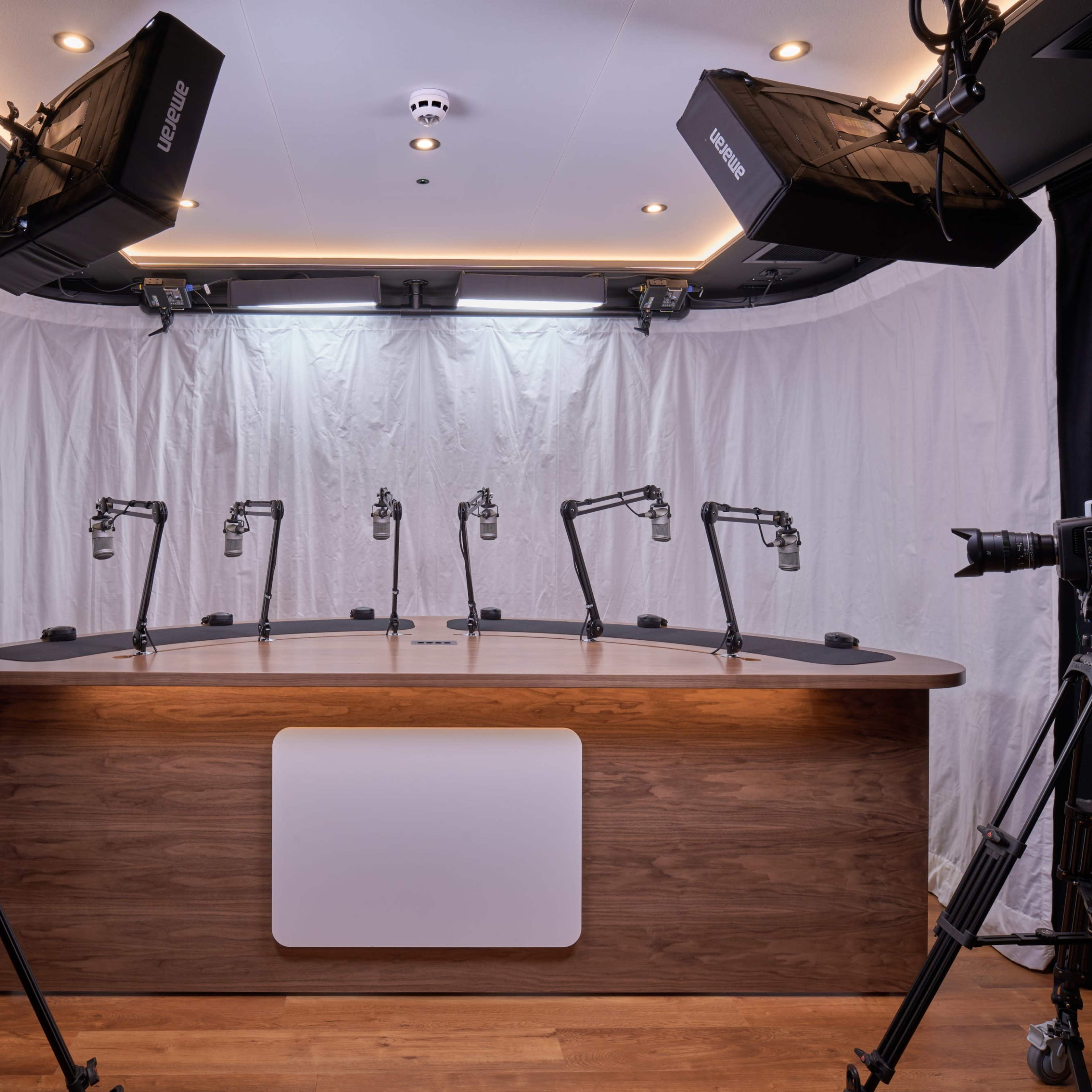 A recording studio setup features a wooden table with six microphone stands and several cameras positioned around it, all surrounded by black curtains and a white backdrop.