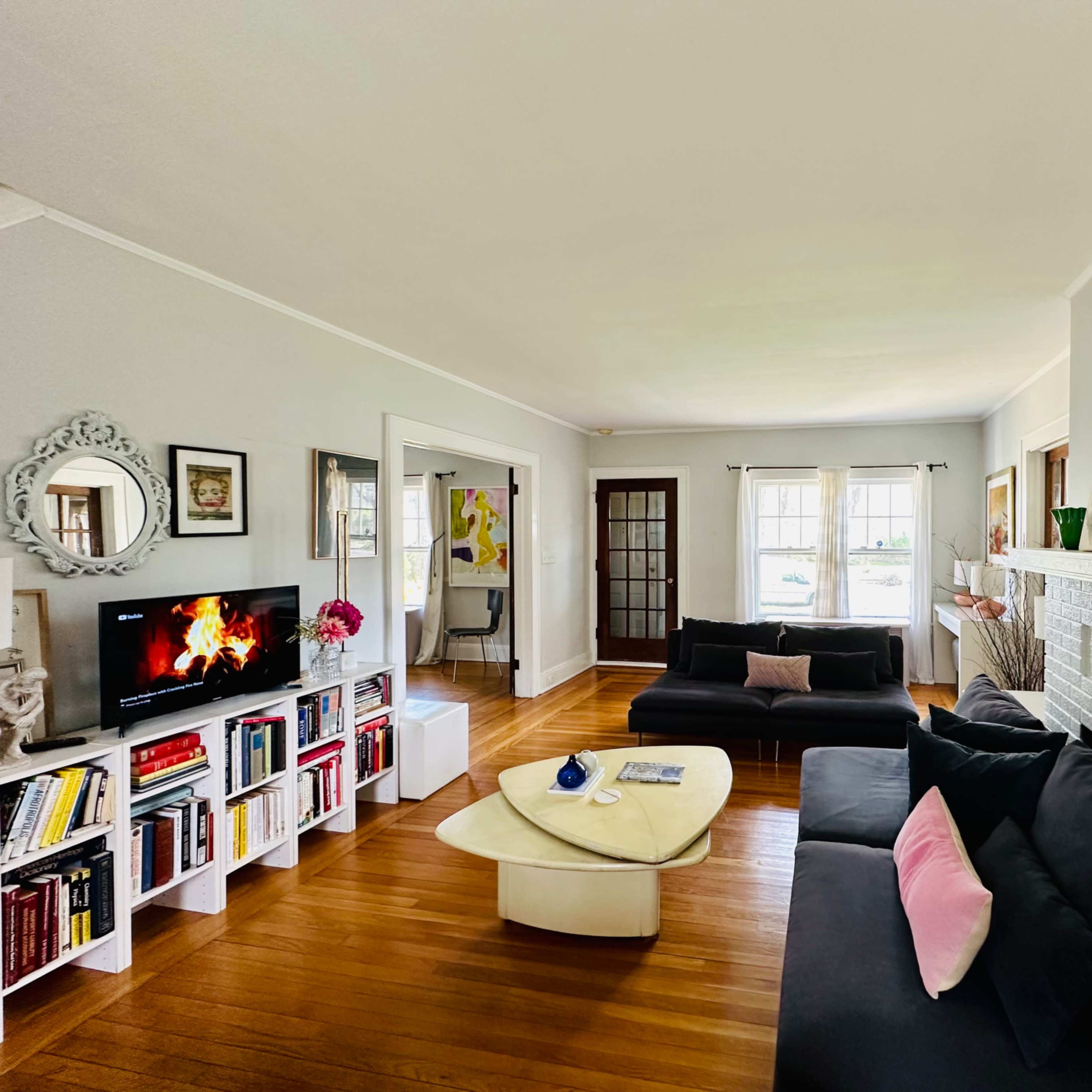 A living room featuring a black sofa, a white coffee table, a bookshelf filled with books, and a television displaying a fireplace scene.