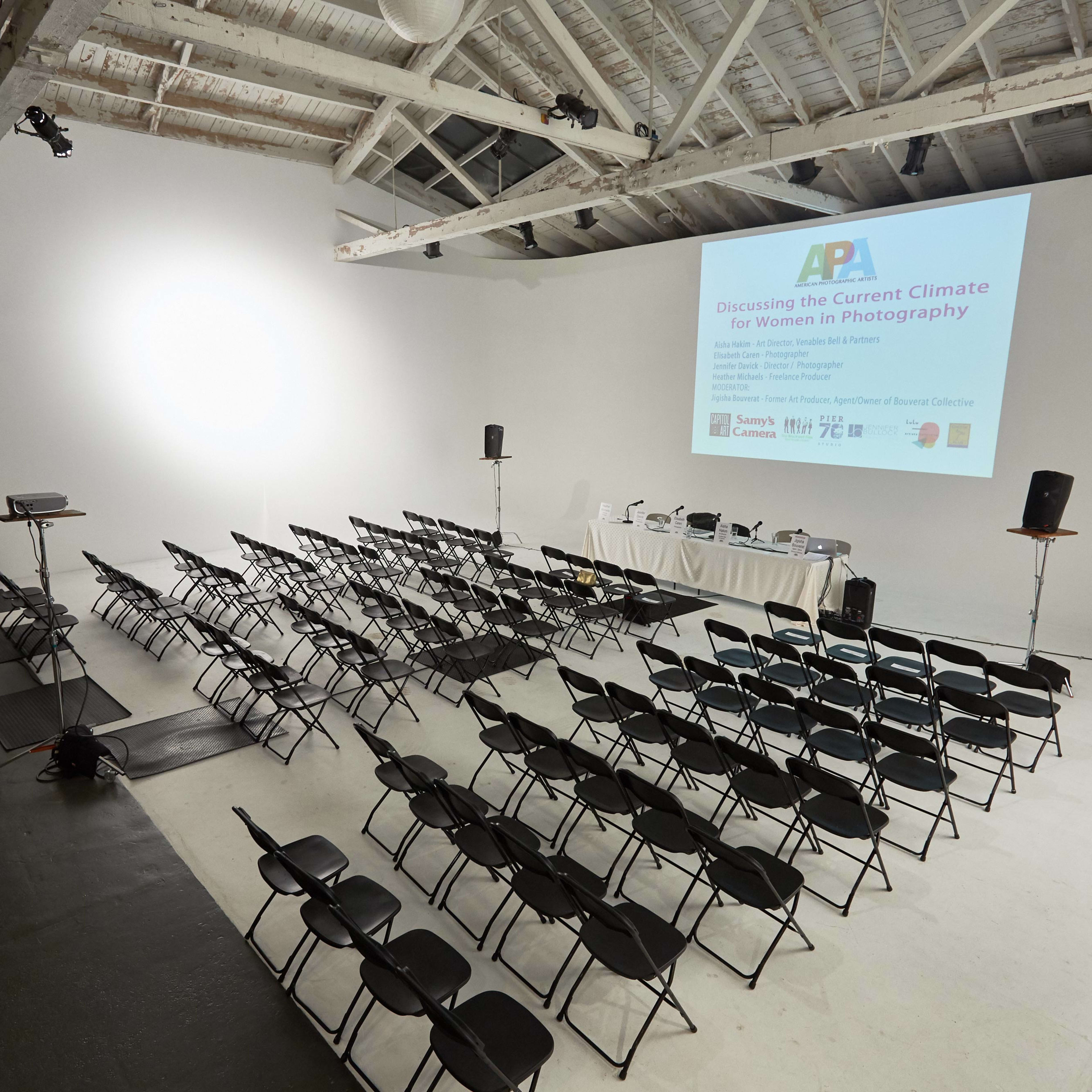 A spacious, well-lit room with rows of black folding chairs arranged facing a presentation screen.