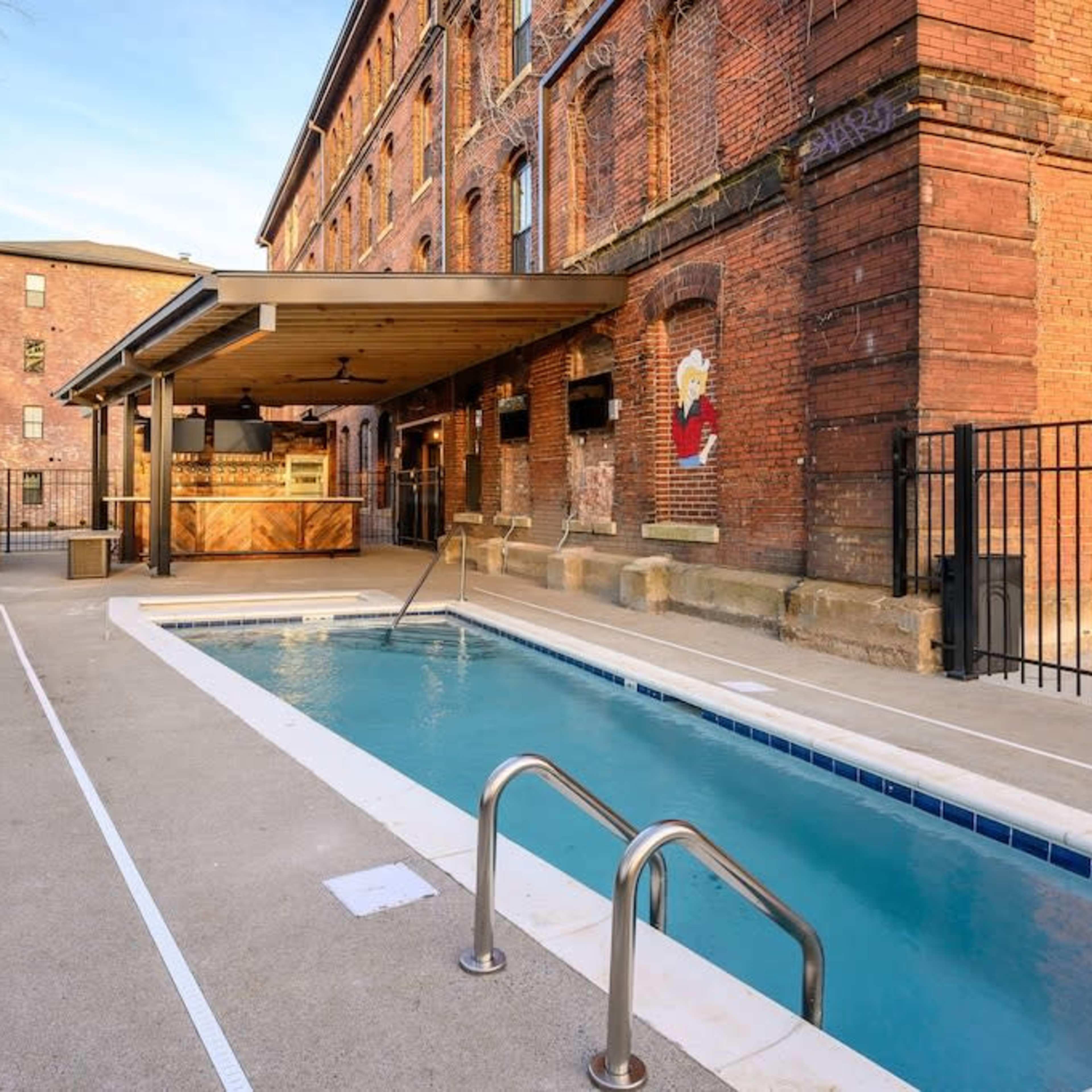 A rectangular swimming pool adjacent to a brick building with a wooden pavilion and a fenced area.