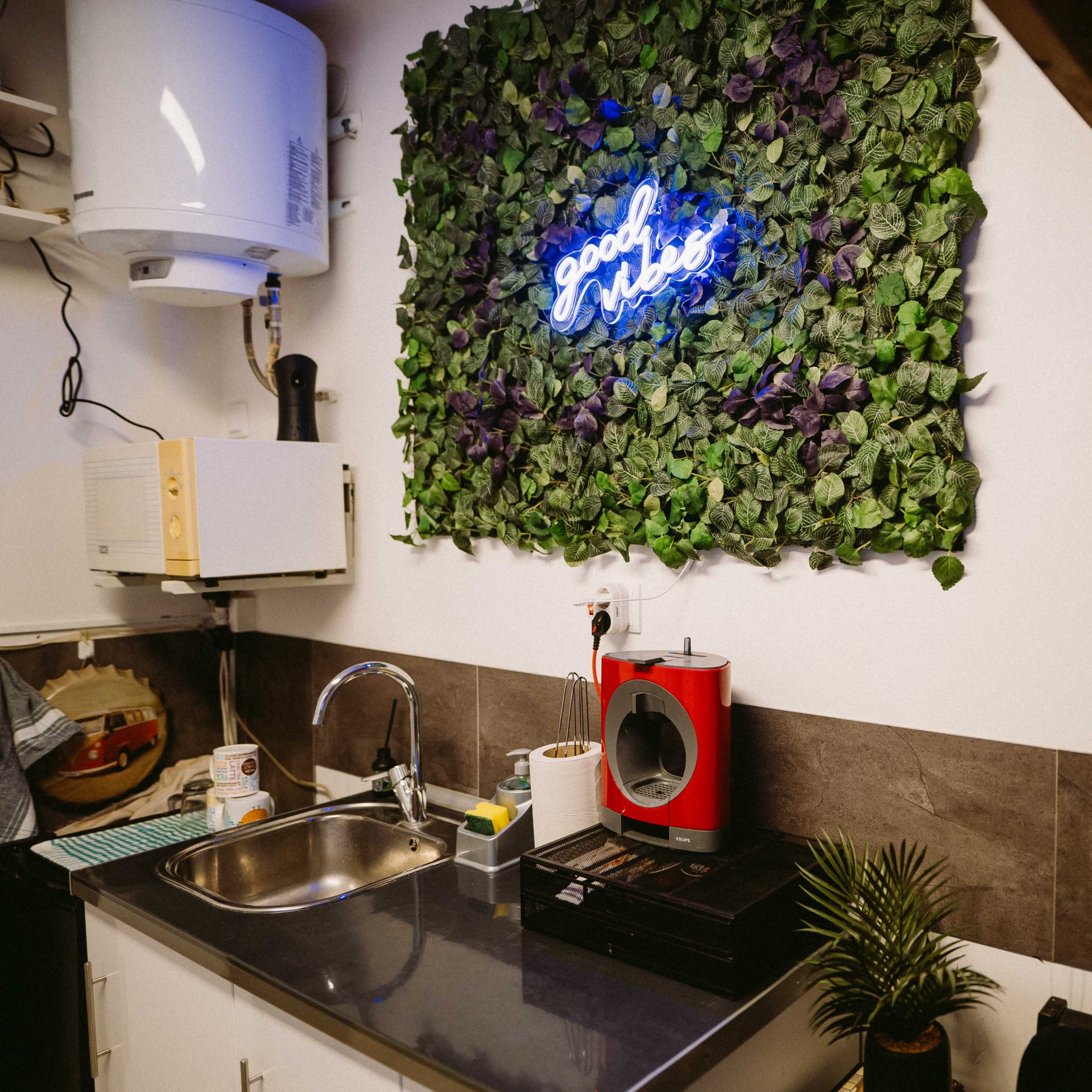 A small kitchen area with a stainless steel sink, a green plant, and a neon sign that reads "good vibes" surrounded by artificial foliage on the wall.