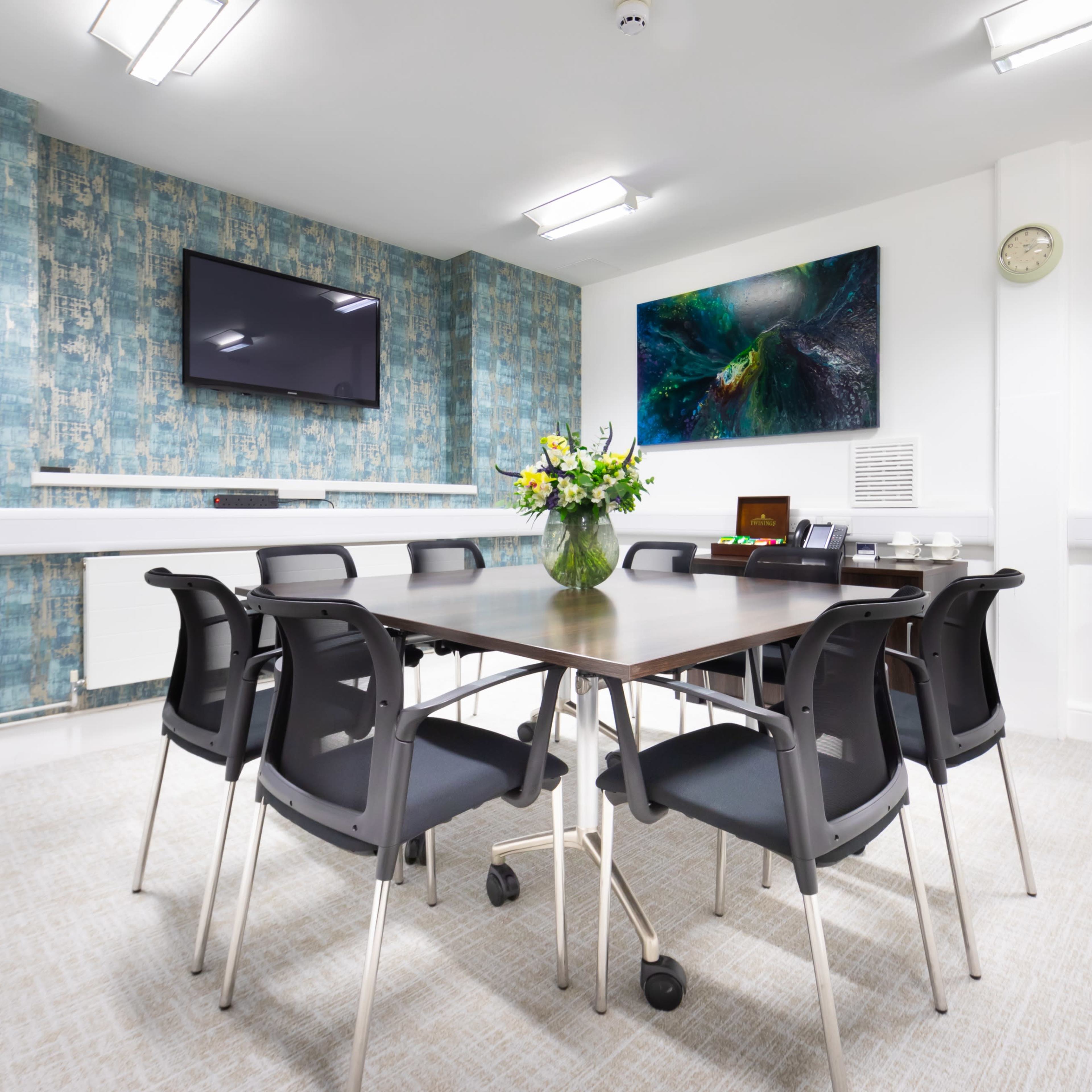 A conference room features a rectangular table surrounded by eight black chairs, with a television on the wall and a vase of flowers as a centerpiece.