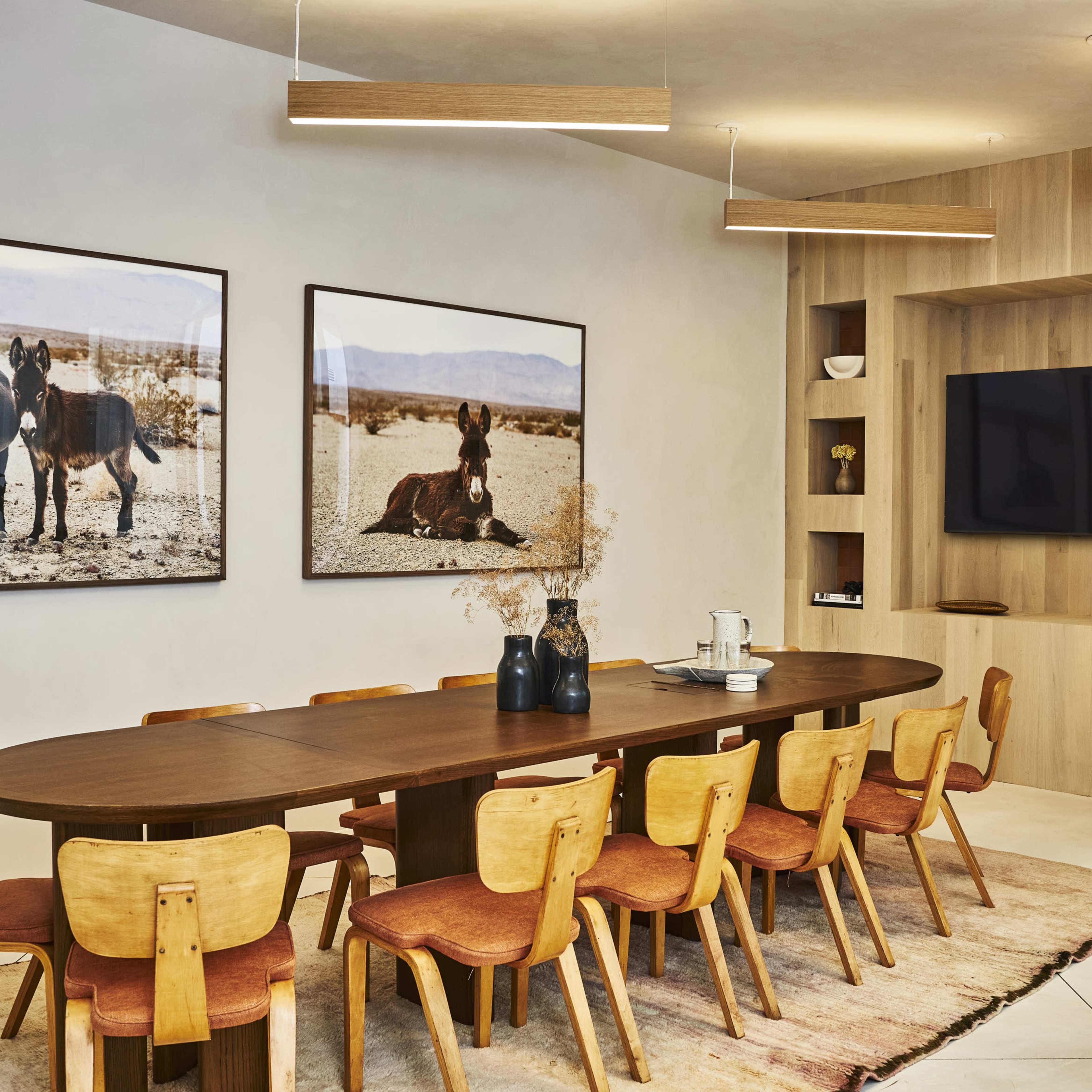A modern dining room features a long wooden table surrounded by chairs, with large photographs of horses on the walls and a TV mounted within a wooden display unit.