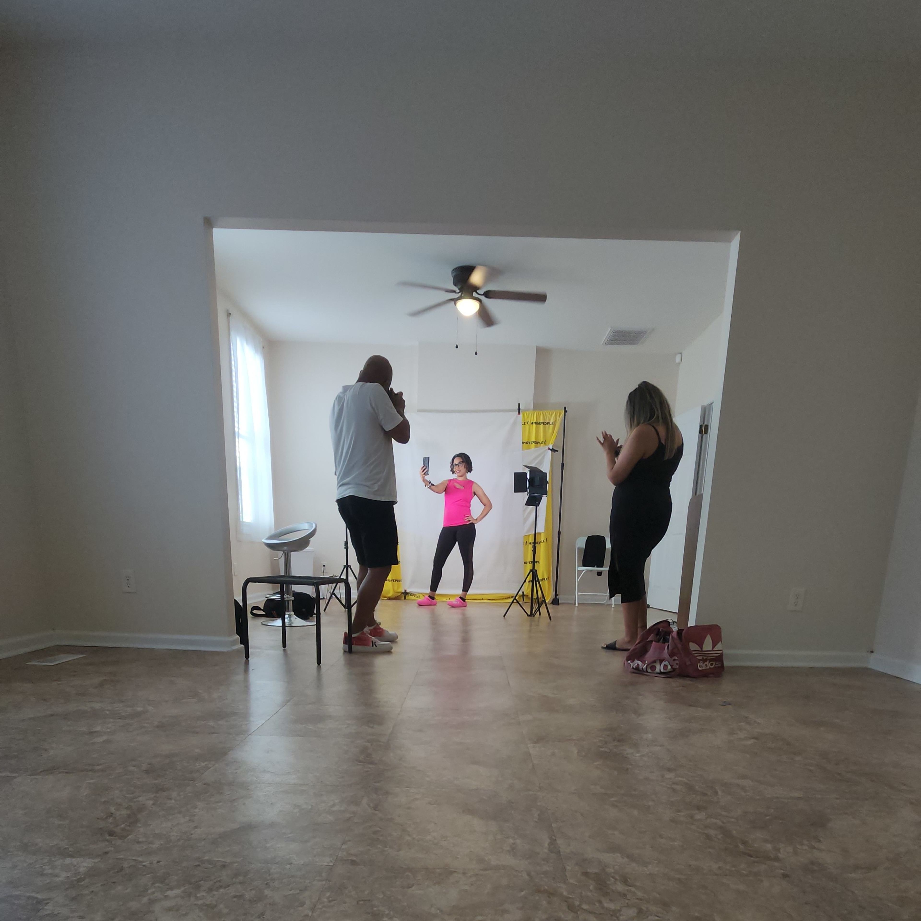 A group of three people is participating in a photo shoot in a spacious, empty room with light-colored walls and a ceiling fan.