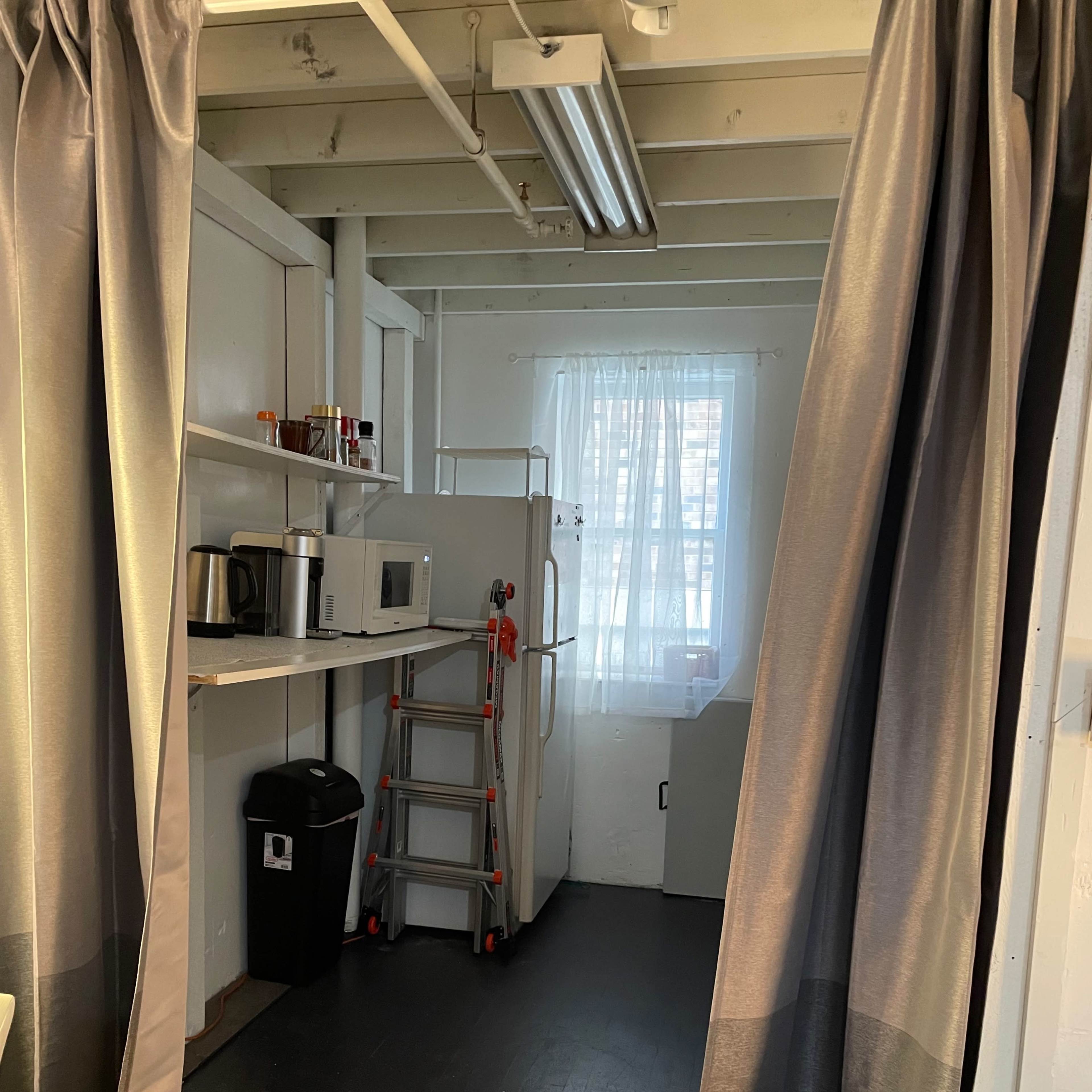 A small kitchen area with cabinets, appliances, a window, and a ladder, divided by gray curtains.