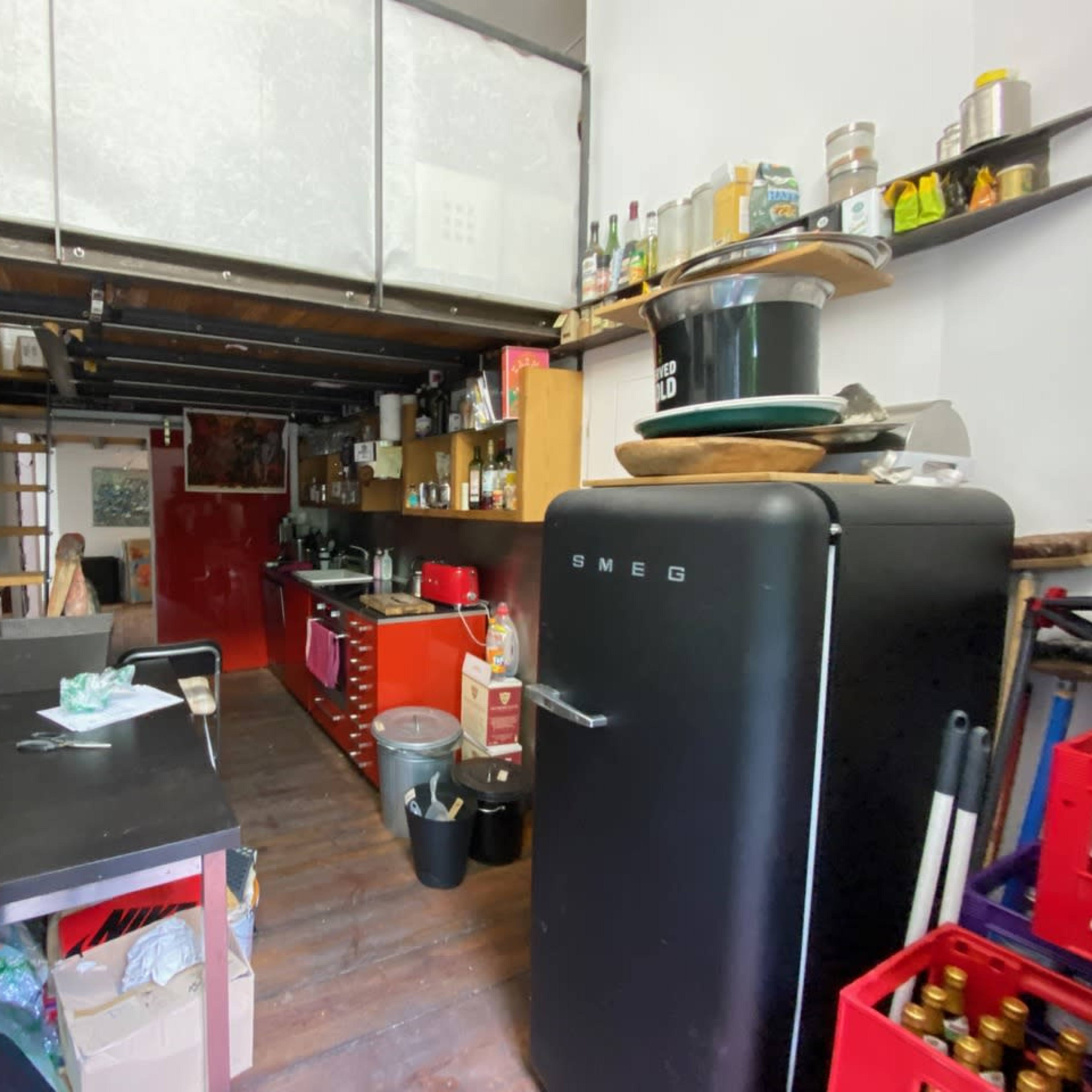 A small kitchen area featuring red cabinets, a vintage-style black refrigerator, and wooden shelving, with various kitchen items and utensils arranged around the space.
