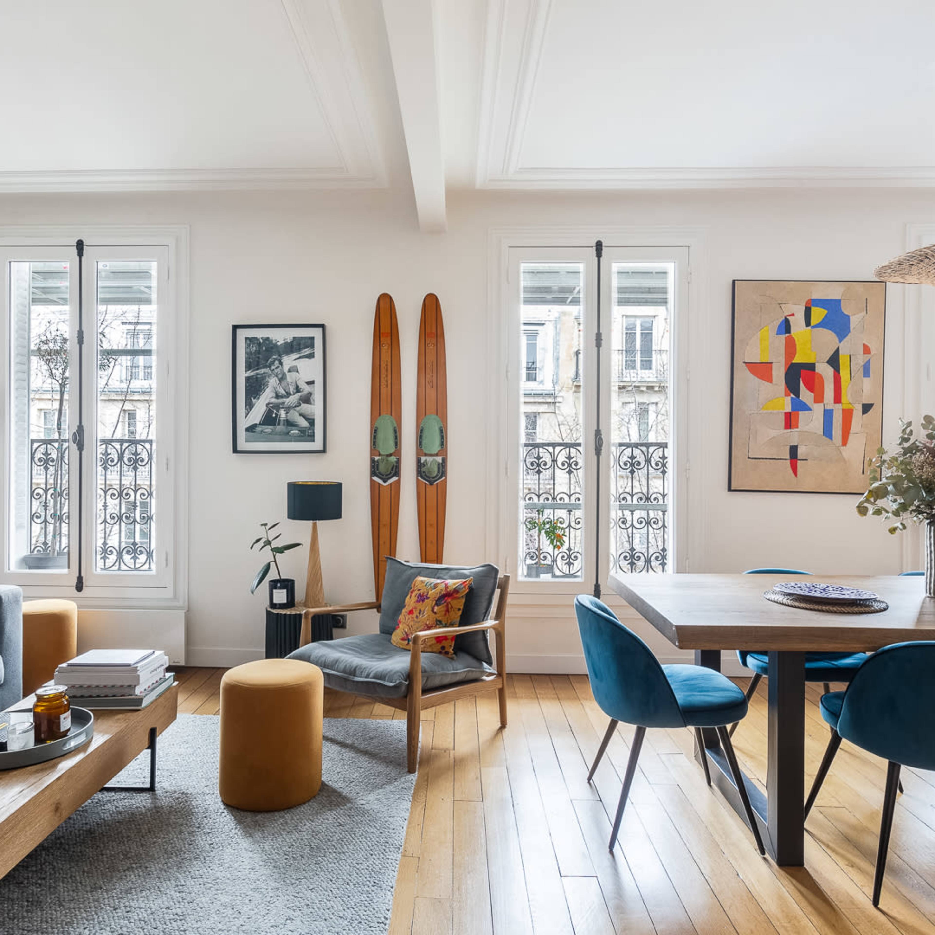 A modern living room with a grey sofa, a wooden coffee table, a dining area with blue chairs, and two decorative wooden skis mounted on the wall.