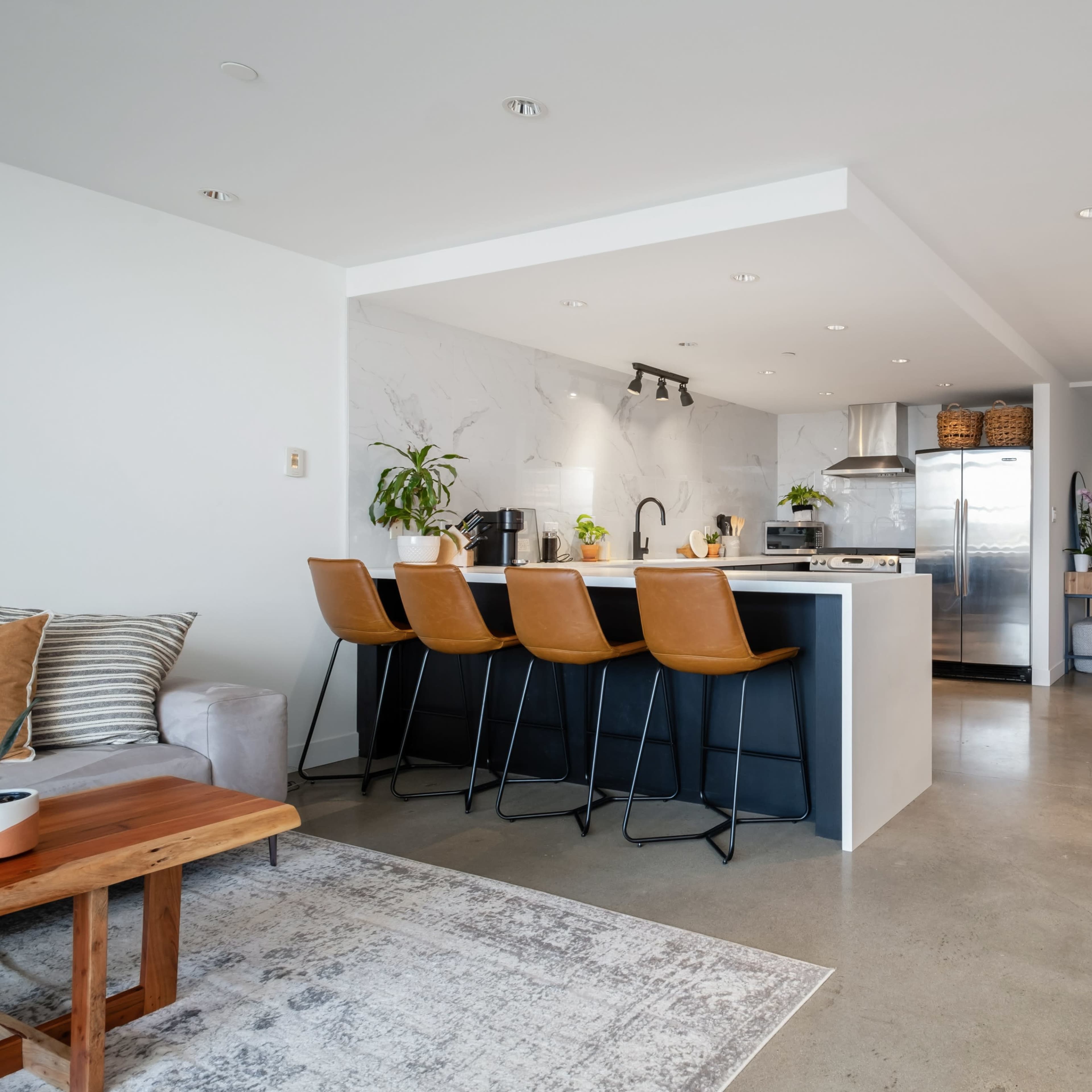 A modern kitchen with a breakfast bar featuring four stools, stainless steel appliances, and a dining area with a black table and wooden chairs.