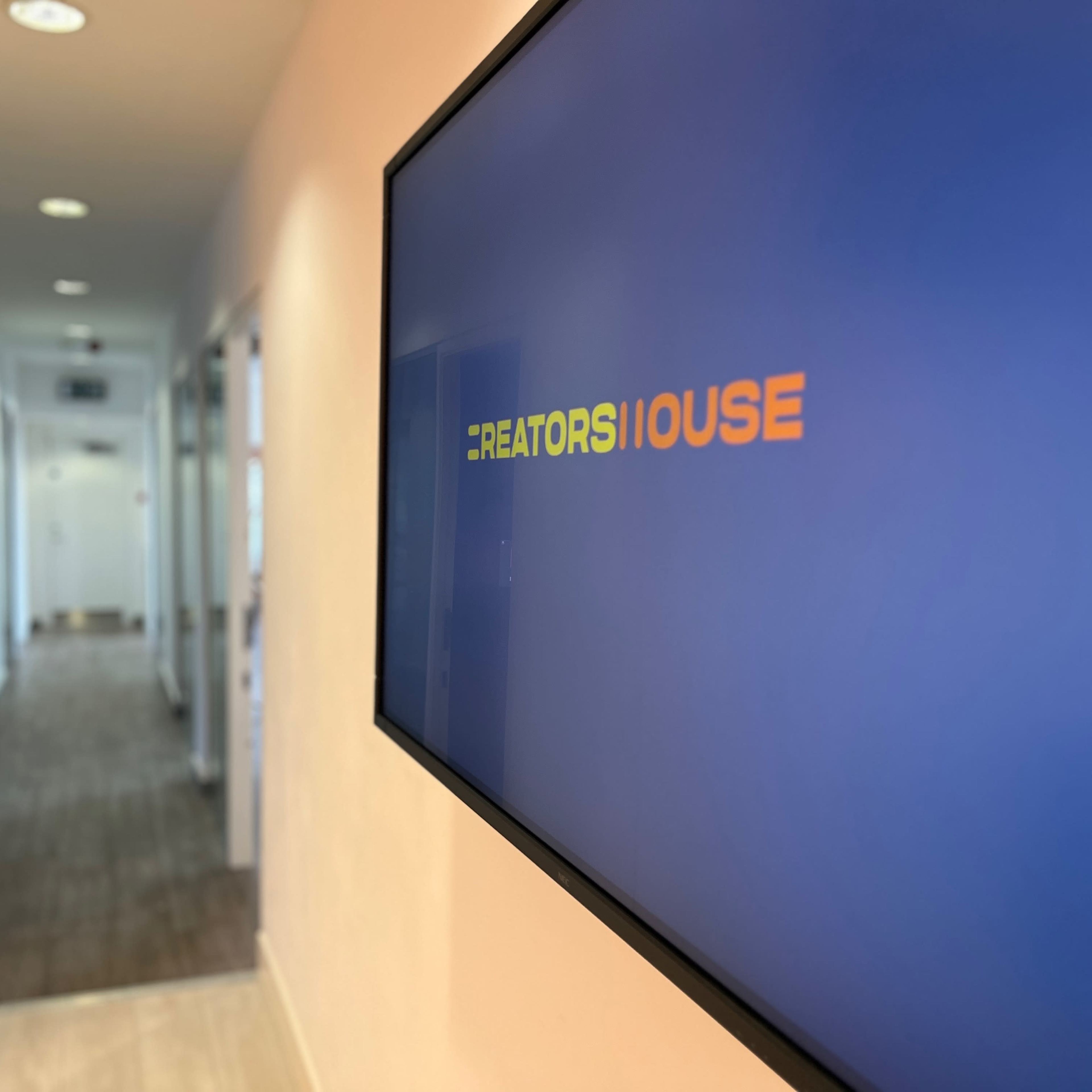 A display screen showing the text "CREATORS HOUSE" is mounted on a wall next to a hallway with several closed office doors.