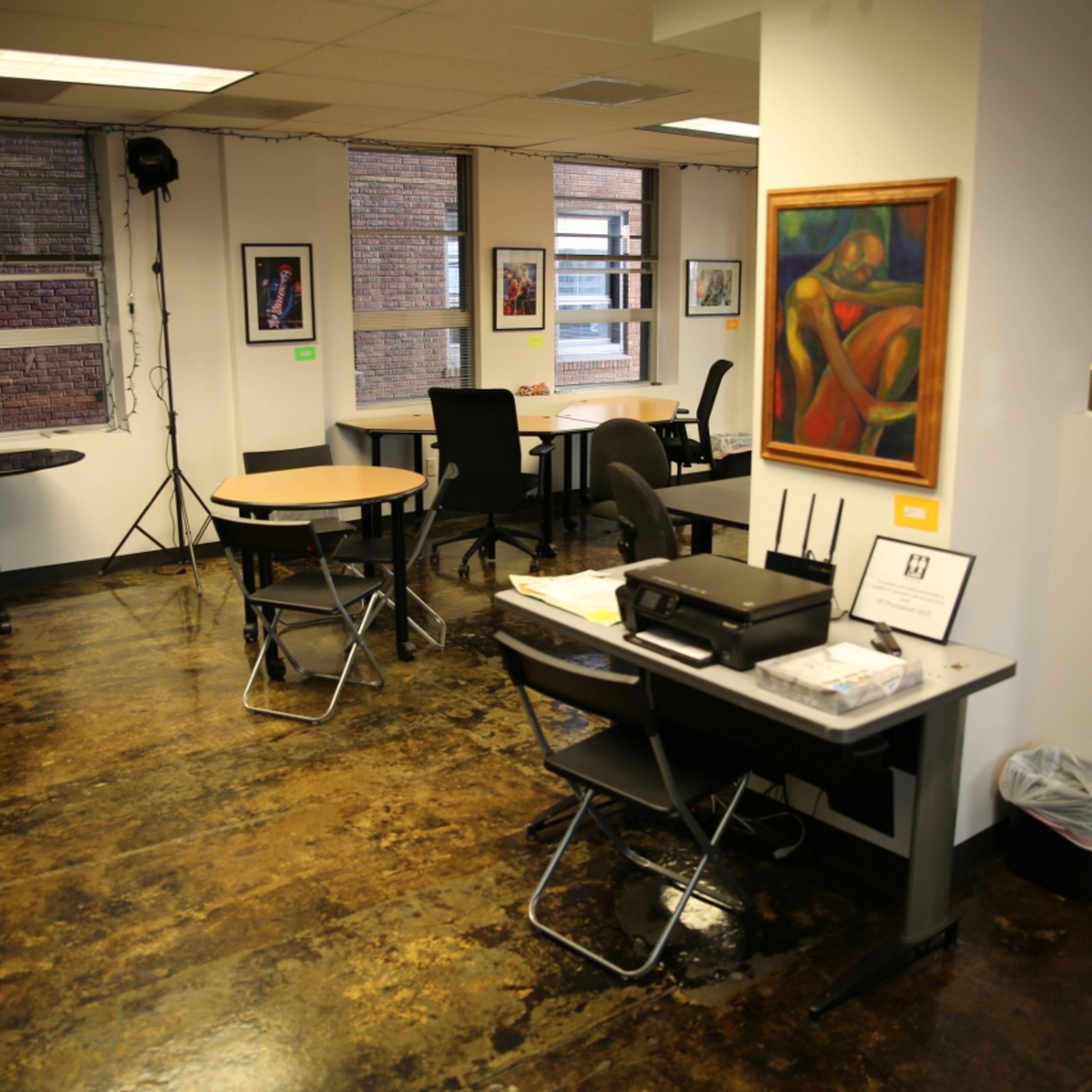 An office space with several tables and chairs, artwork on the walls, and a printer on a desk.