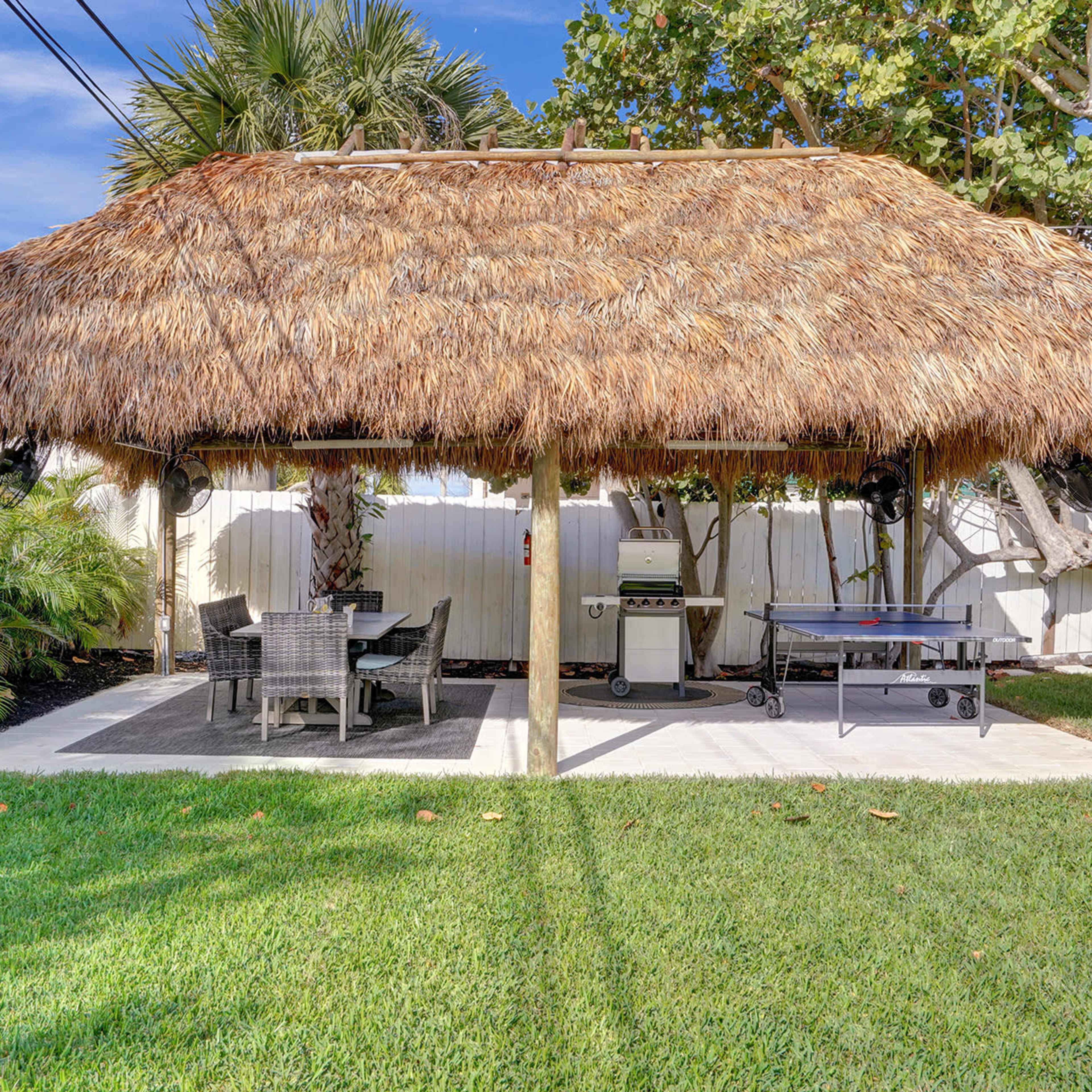 A thatched roof gazebo with a dining area and a grill is situated on a grassy lawn surrounded by palm trees.