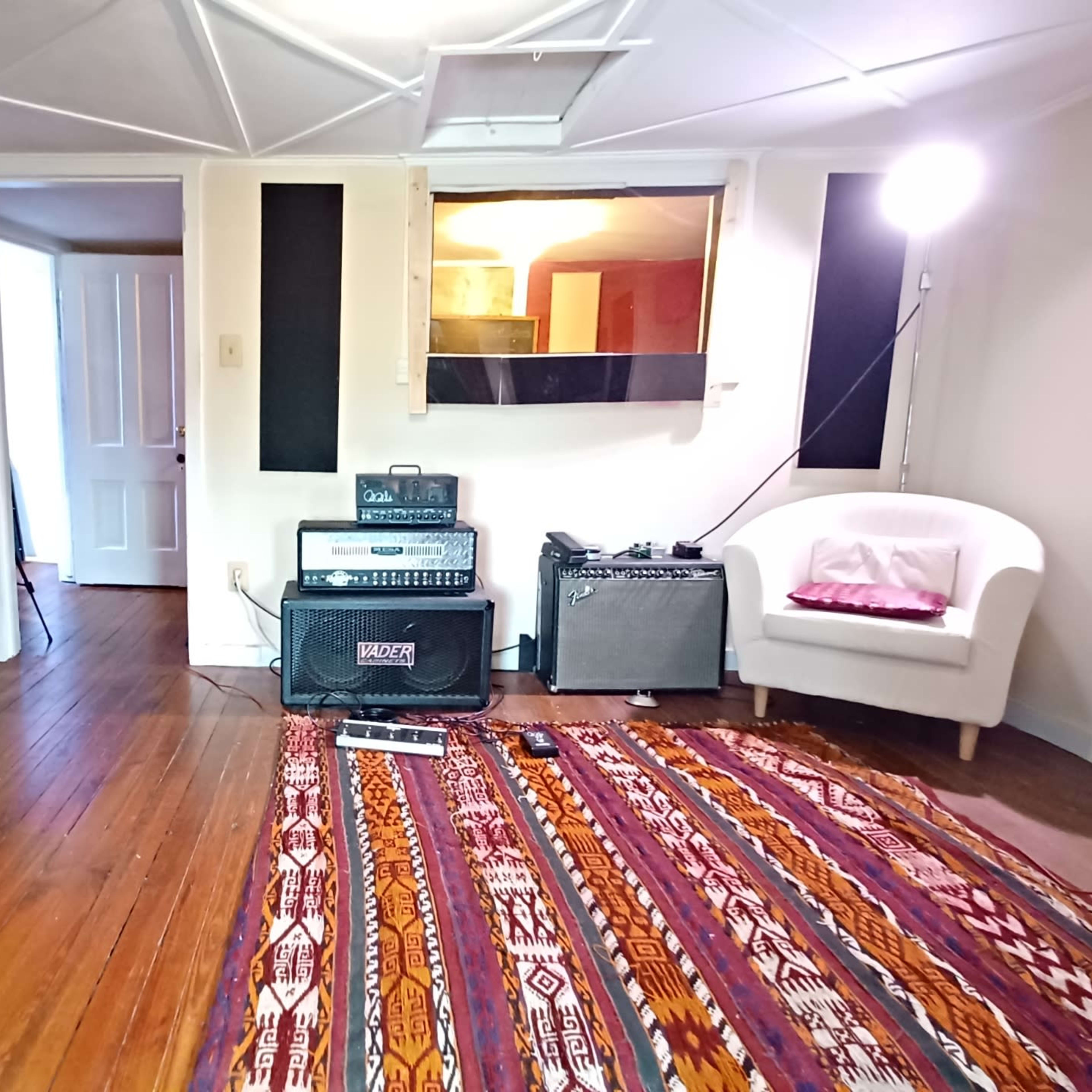 A small music studio with wooden floors, a patterned rug, an amplifier, a guitar stand, and a white chair in a corner.