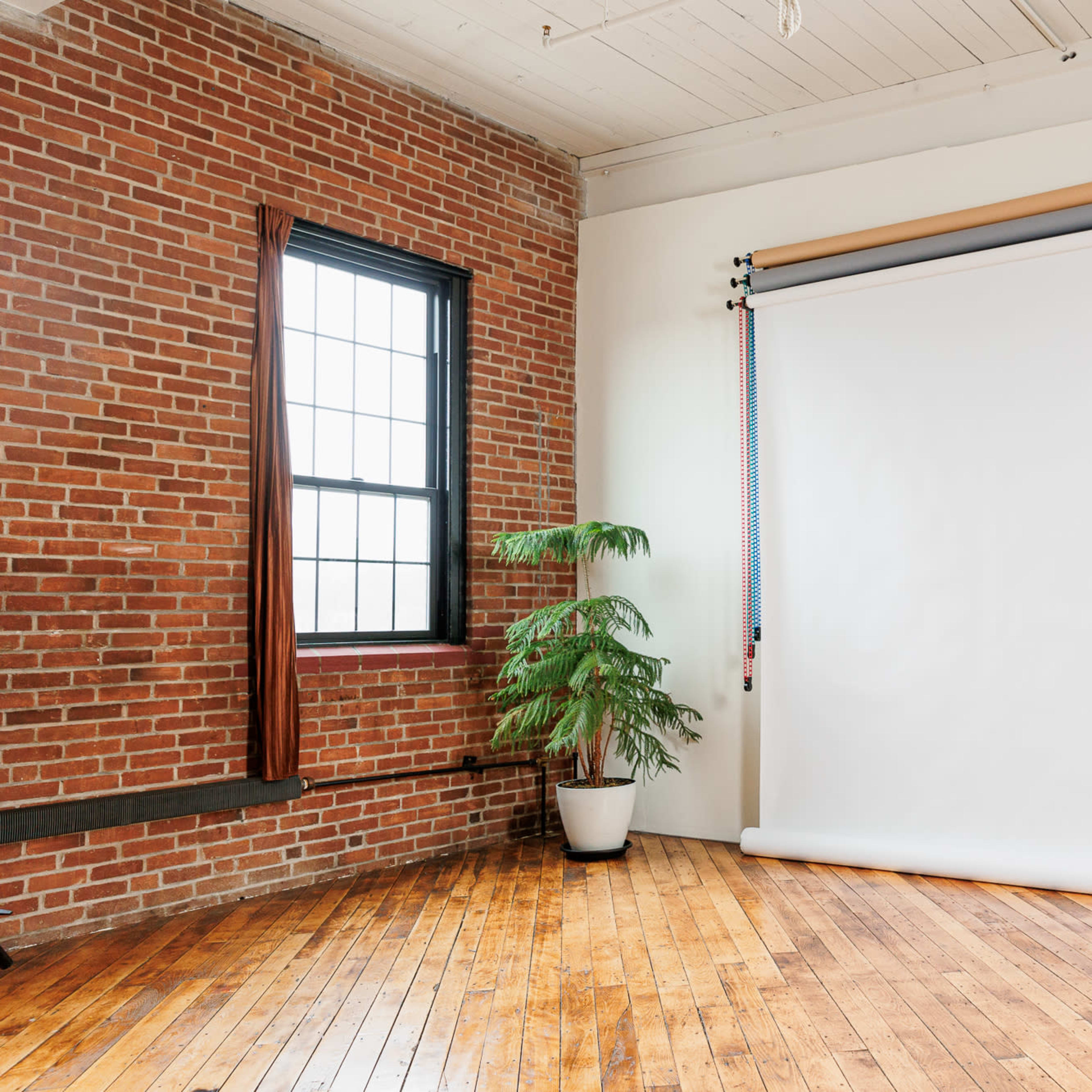 A spacious room with exposed brick walls, a white backdrop on rollers, a potted plant, and a wooden stool on a hardwood floor.