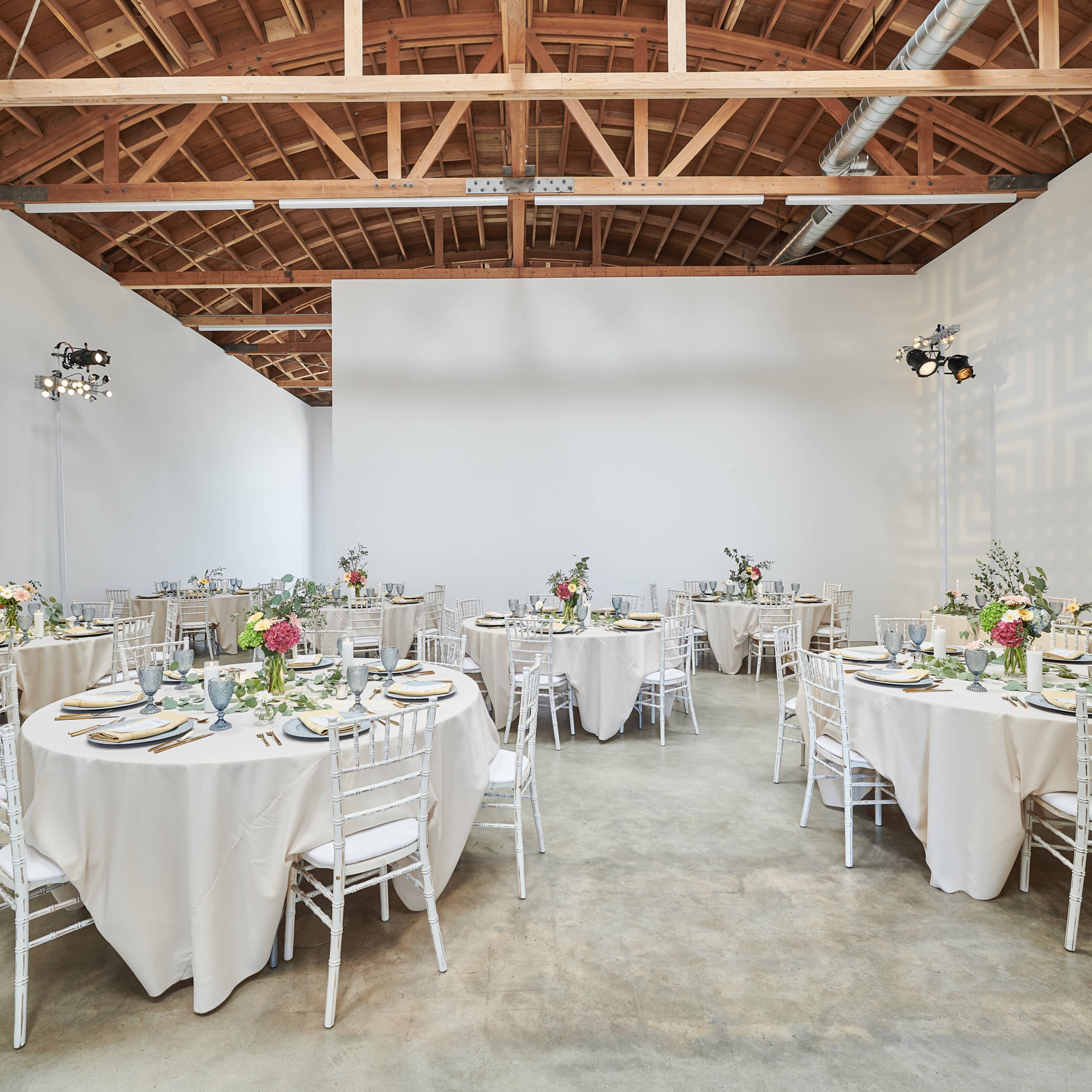 A spacious event room features several round tables set with tablecloths, floral centerpieces, and dining ware.