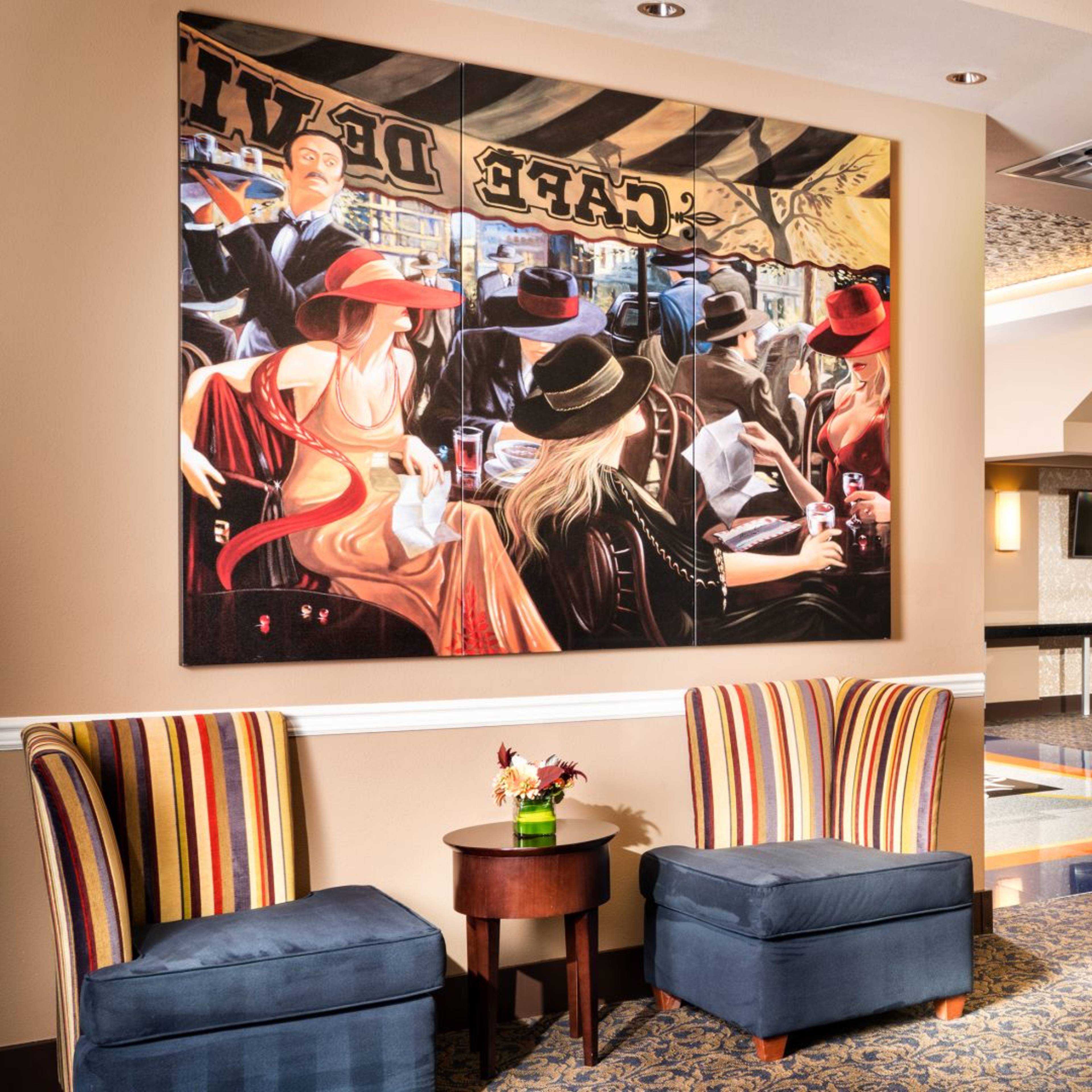 A hotel lobby featuring two striped chairs, a small table with flowers, a large painting on the wall, and a staircase leading to an upper level.