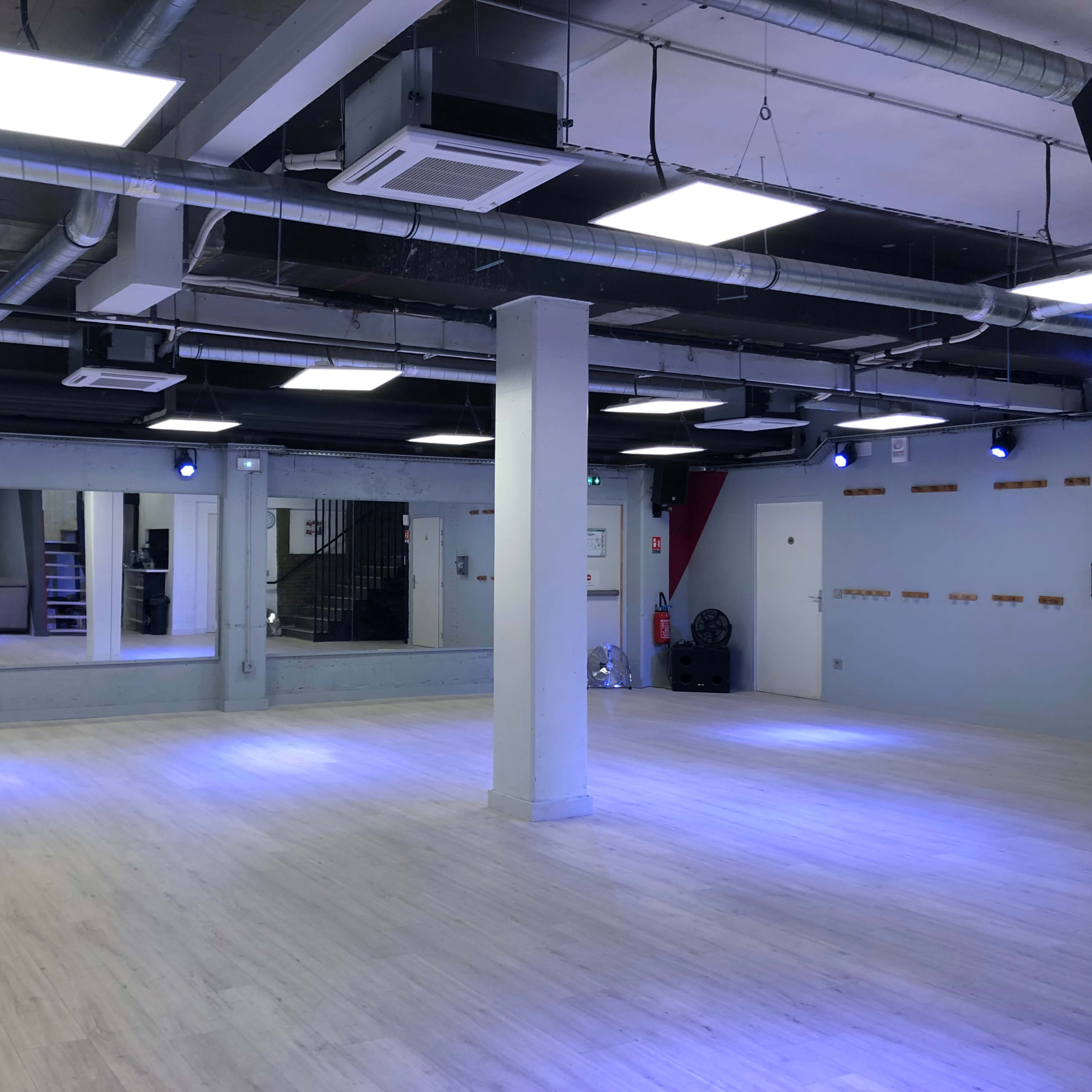 A spacious dance studio with wood flooring, bright ceiling lights, and large mirrors along one wall.