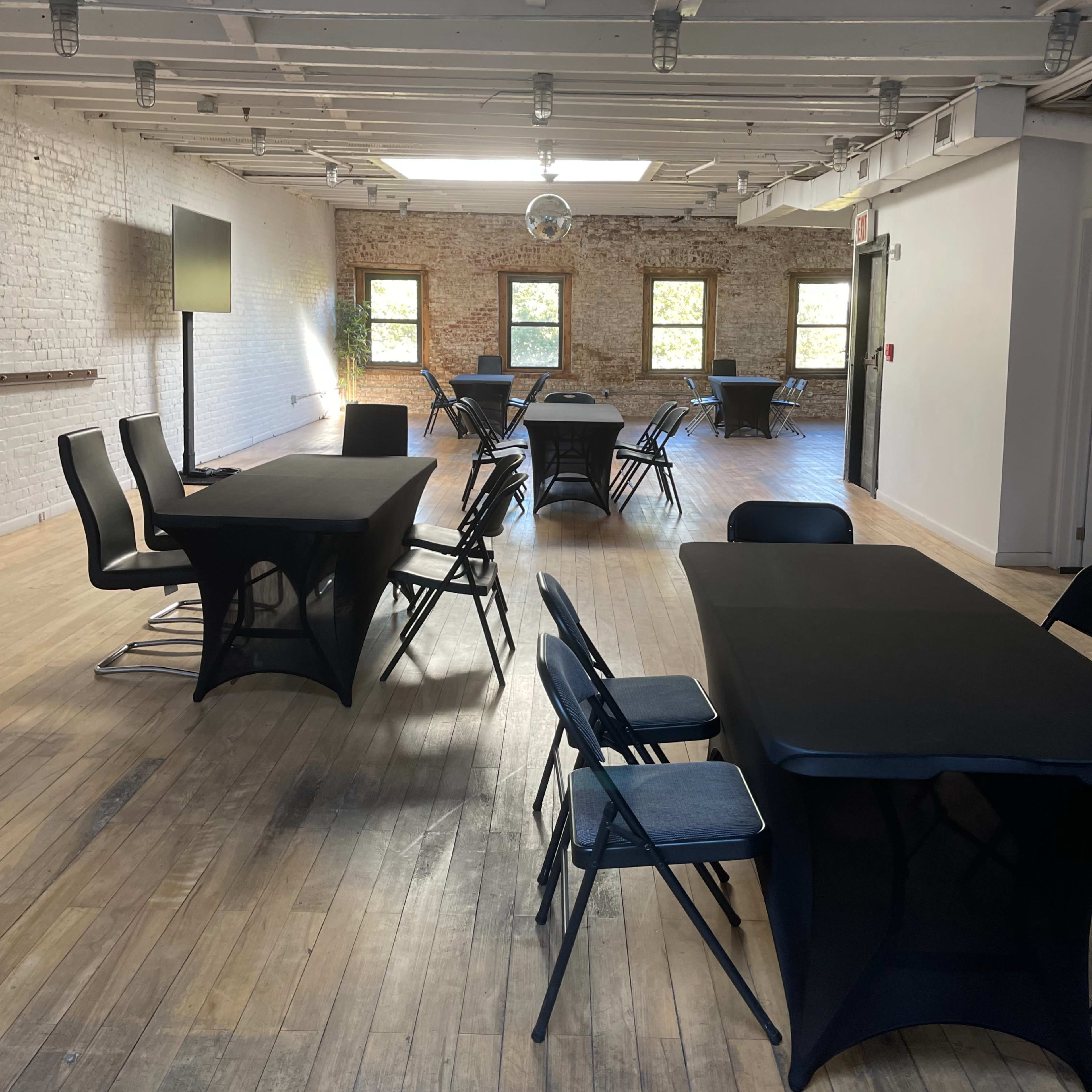 A spacious room with wooden floors, featuring several black tables and chairs arranged throughout, and large windows allowing natural light to enter.
