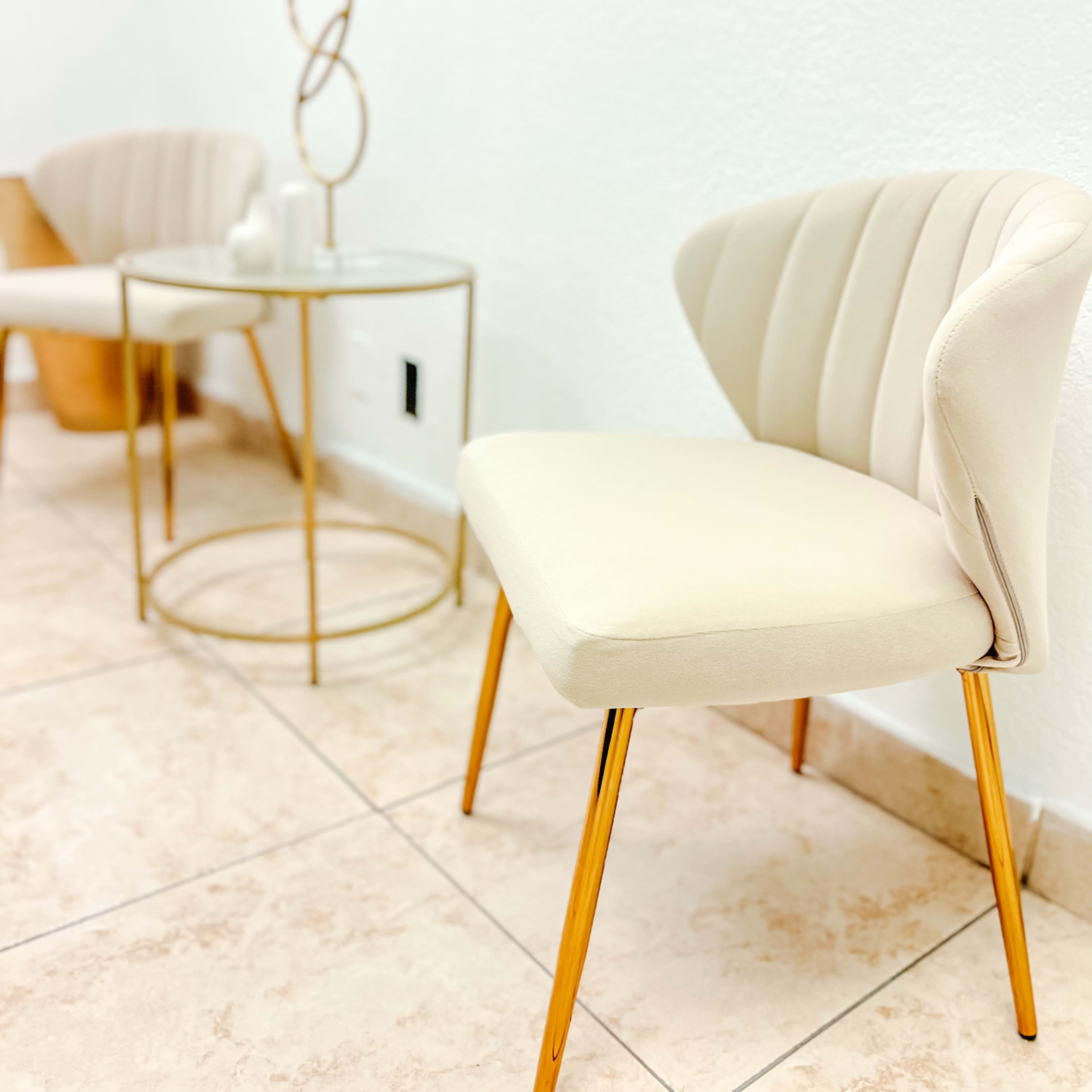 A light-colored, upholstered chair with gold legs sits beside a circular gold side table in a room with tiled flooring and white walls.